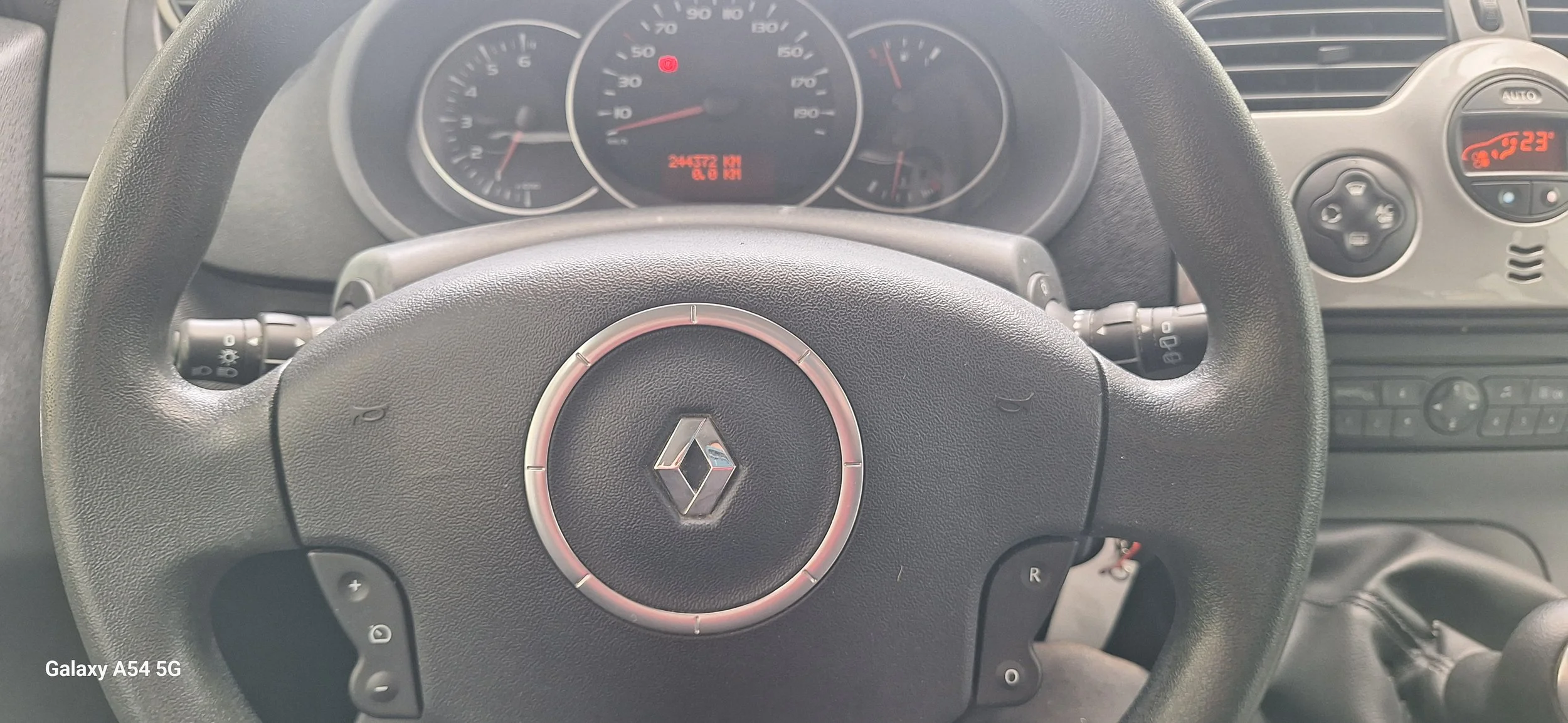Inside a vehicle, view of the black steering wheel with a Renault logo, dashboard with speedometer, tachometer, fuel gauge, and a small digital display showing temperature and other indicators. To the right, part of the climate control system and center console are visible.