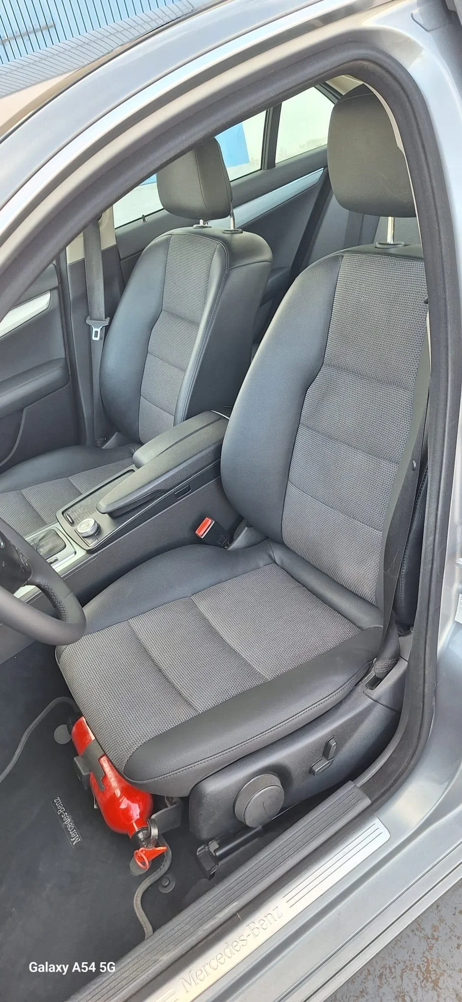 Inside a vehicle, view of the black steering wheel with a Renault logo, dashboard with speedometer, tachometer, fuel gauge, and a small digital display showing temperature and other indicators. To the right, part of the climate control system and center console are visible.