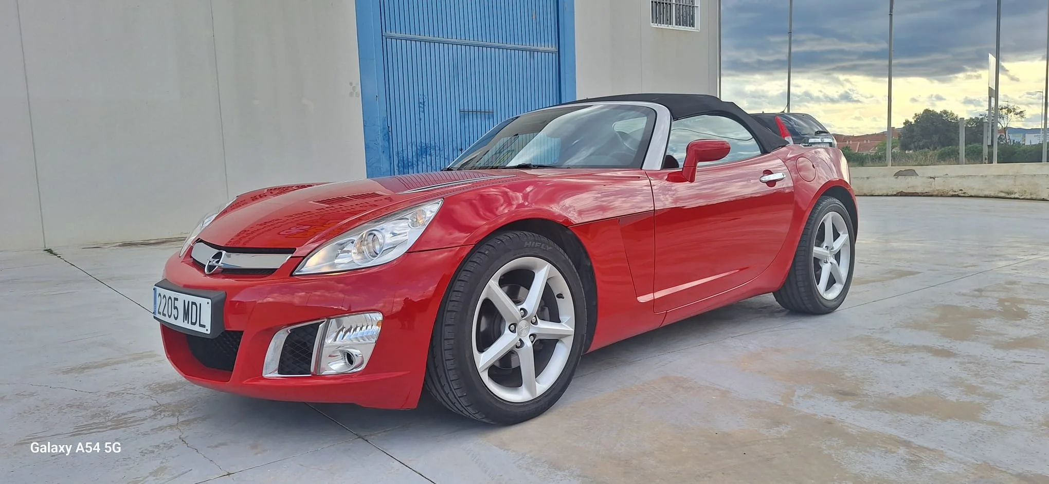 The open rear of a minivan showing the cargo area with a blue item on the left side and the back seats in an upright position. The minivan is parked on a concrete surface with a beige wall and a large planter with green plants in the background.