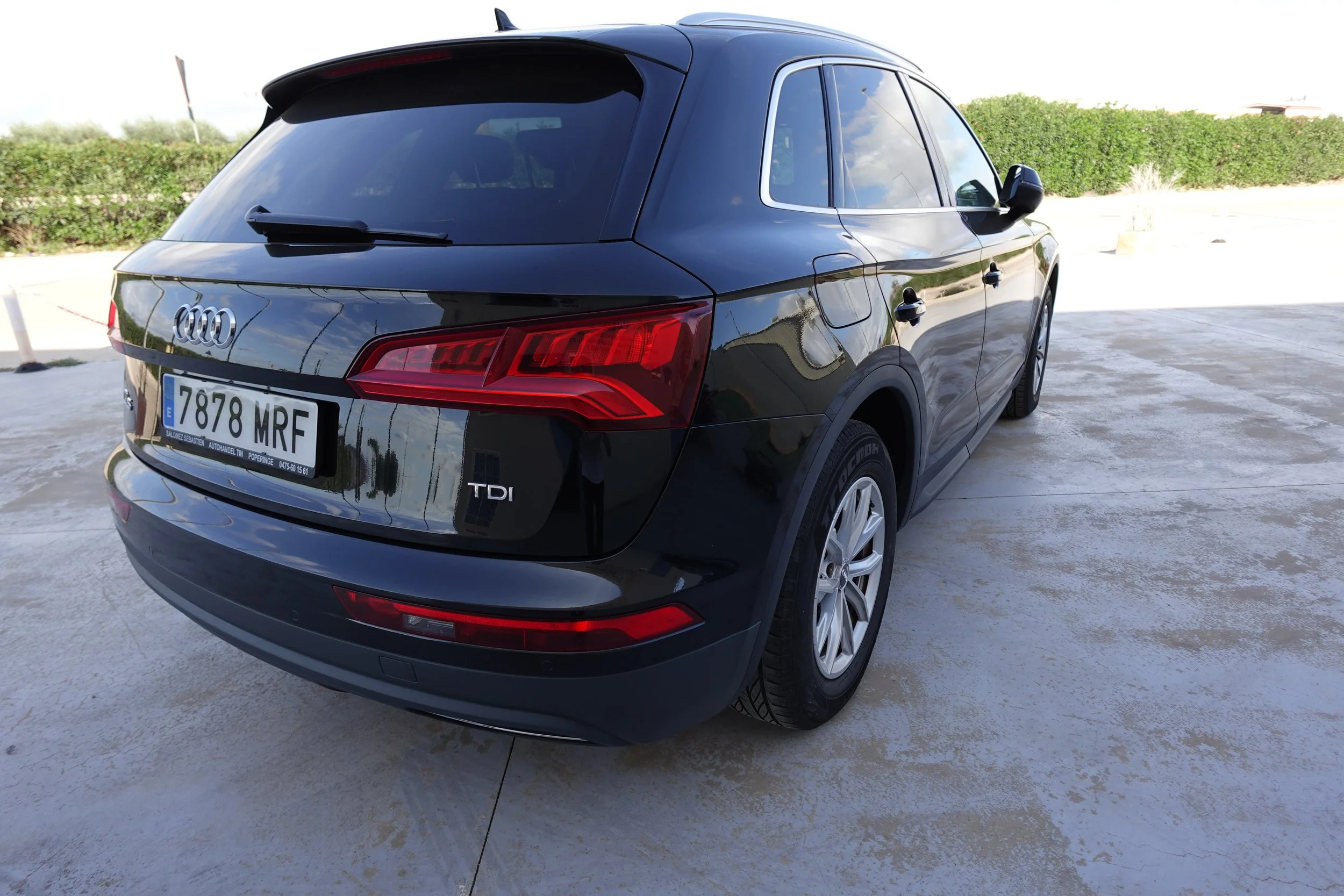 Black Audi SUV parked on a concrete surface, with a background of green bushes and a partly cloudy sky.