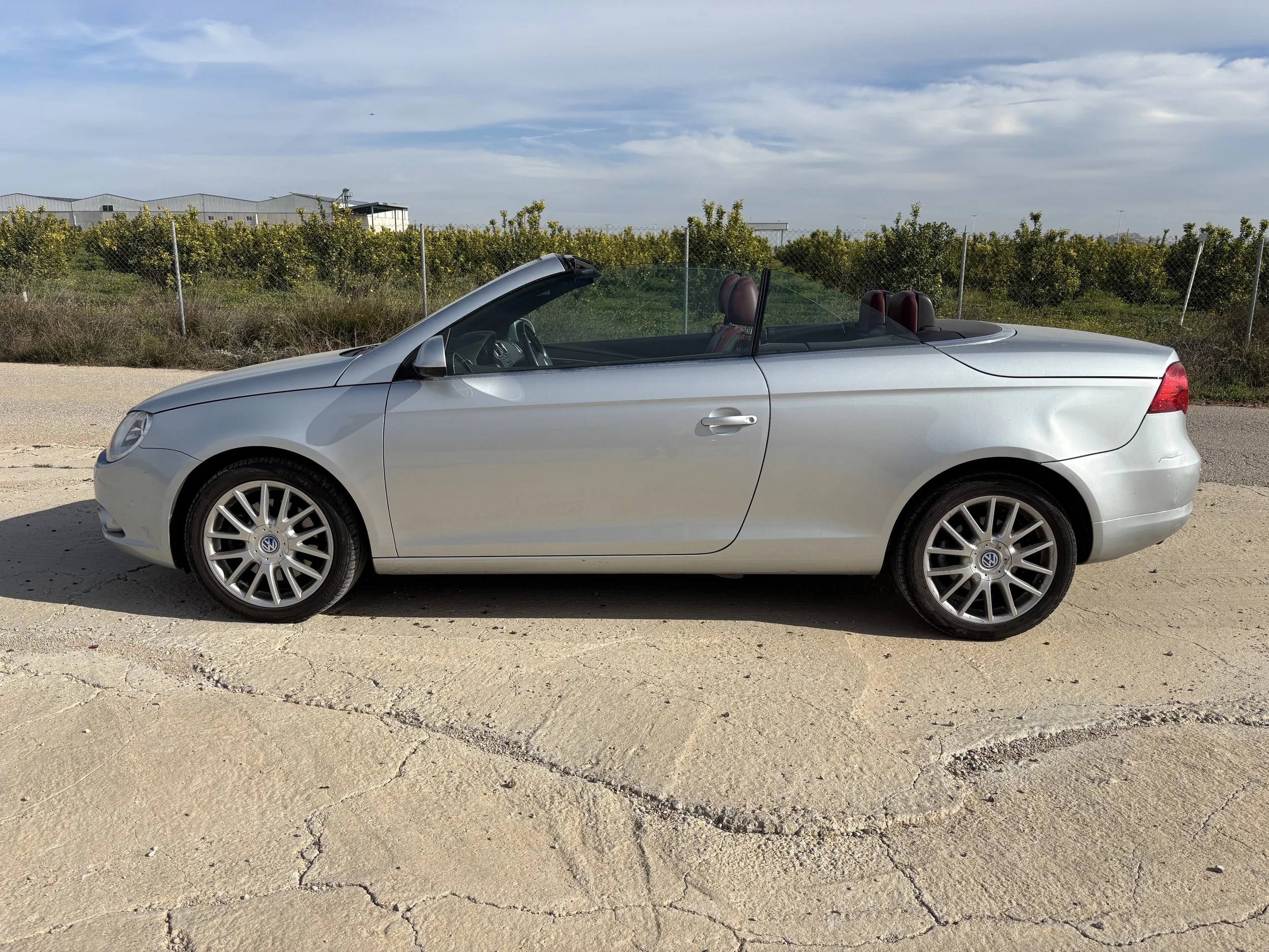 The open rear of a minivan showing the cargo area with a blue item on the left side and the back seats in an upright position. The minivan is parked on a concrete surface with a beige wall and a large planter with green plants in the background.