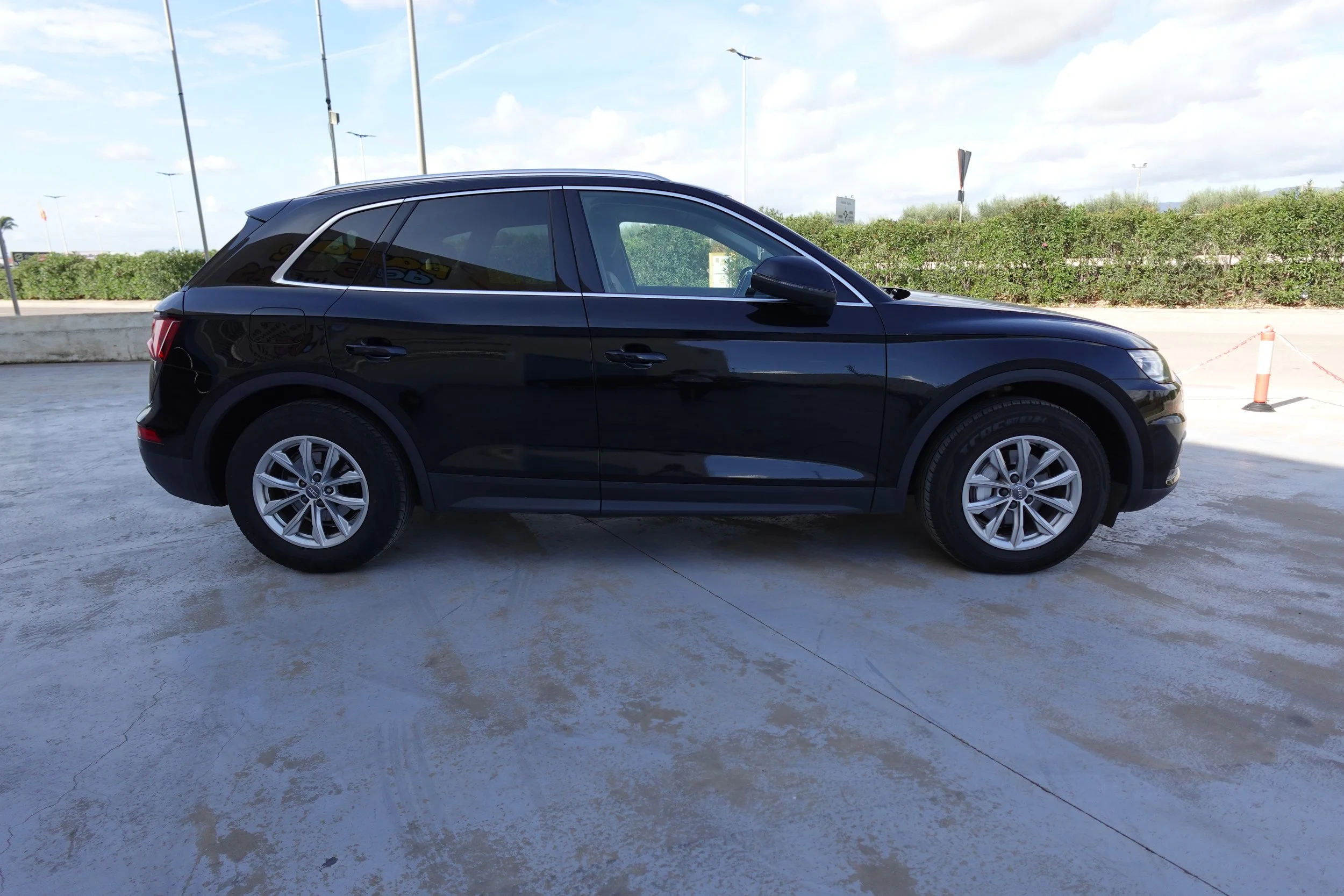 Side view of a black SUV parked outdoors on a concrete surface with a hedge, street signs, and a partly cloudy sky in the background.