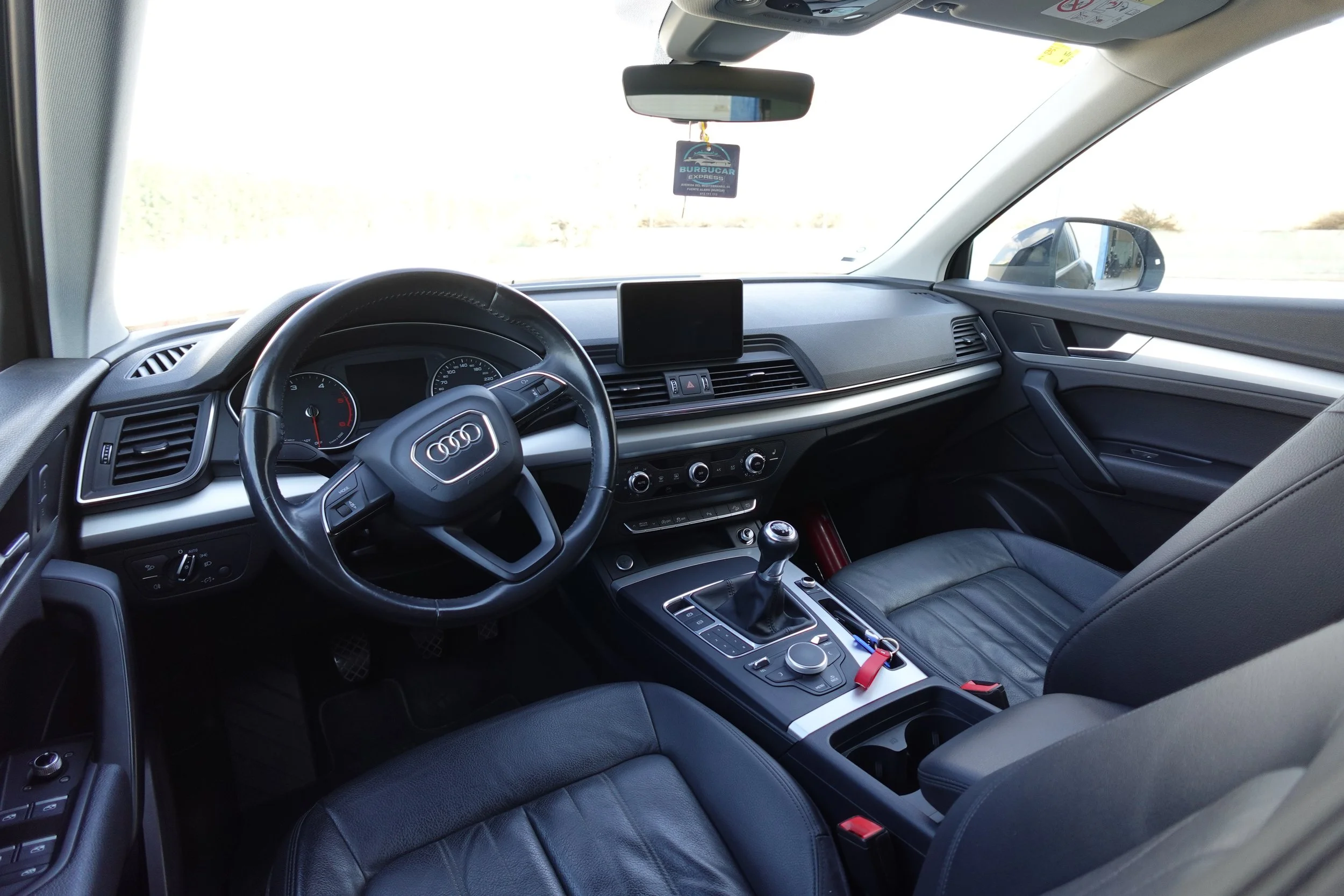 Interior view of an Audi vehicle, showing the steering wheel, dashboard, gear shift, and front seats with a view of the road outside.