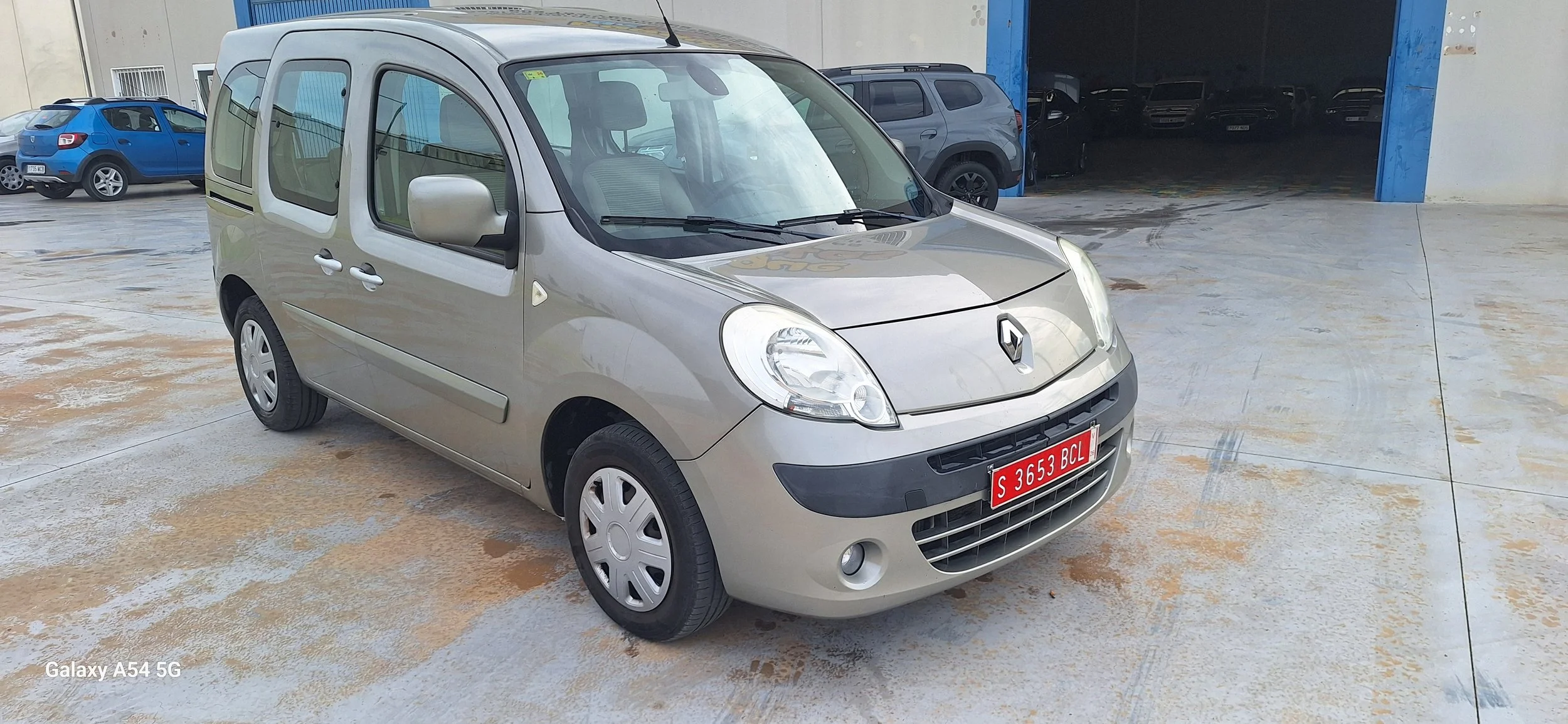 A beige Renault compact car with a red license plate parked on a wet concrete surface outside a building, with other vehicles and a blue garage door in the background.