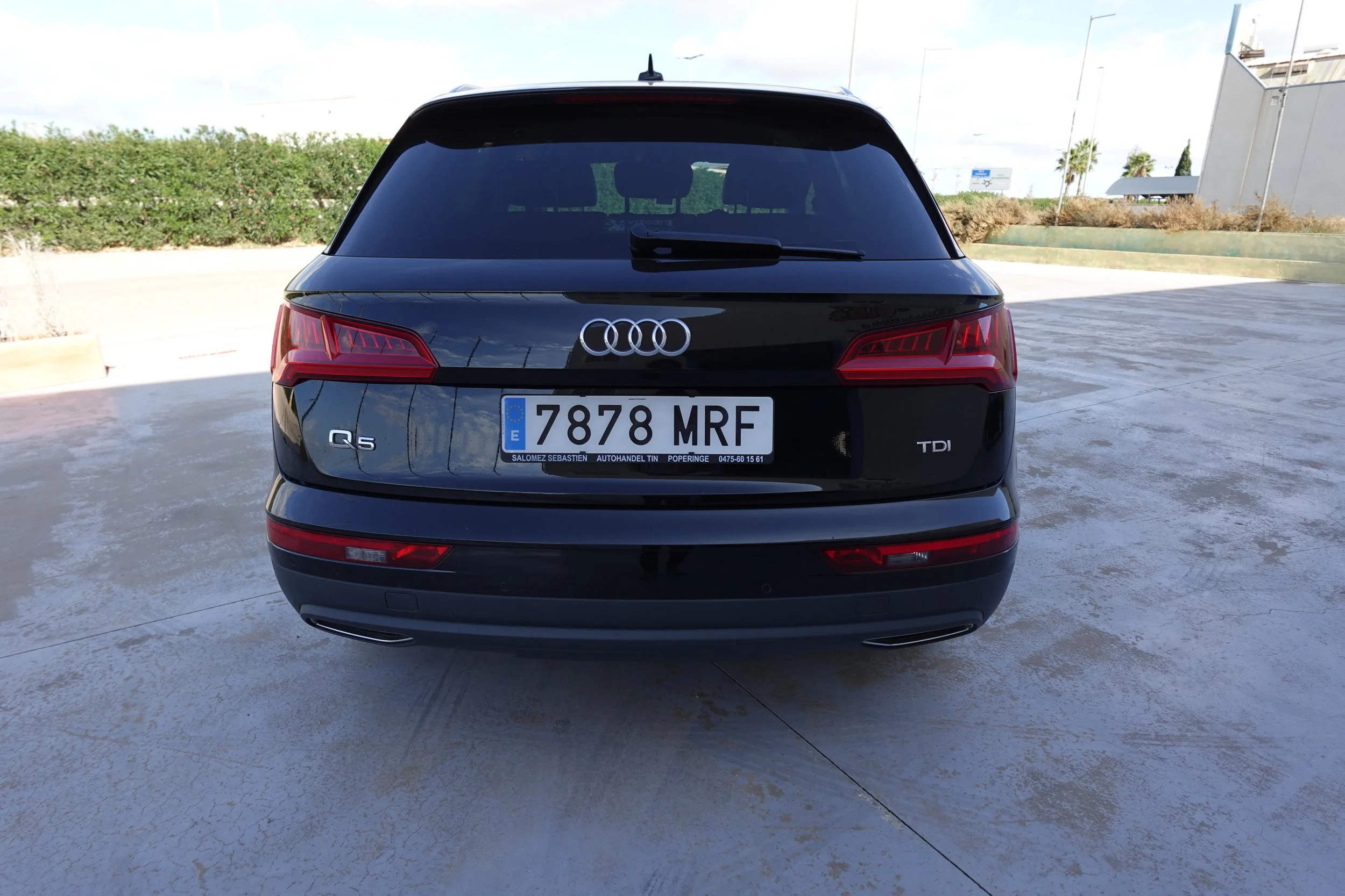 Rear view of a black Audi Q5 SUV with a European license plate, parked on a concrete surface with green bushes and a partly cloudy sky in the background.