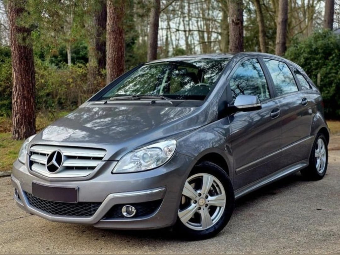 The open rear of a minivan showing the cargo area with a blue item on the left side and the back seats in an upright position. The minivan is parked on a concrete surface with a beige wall and a large planter with green plants in the background.