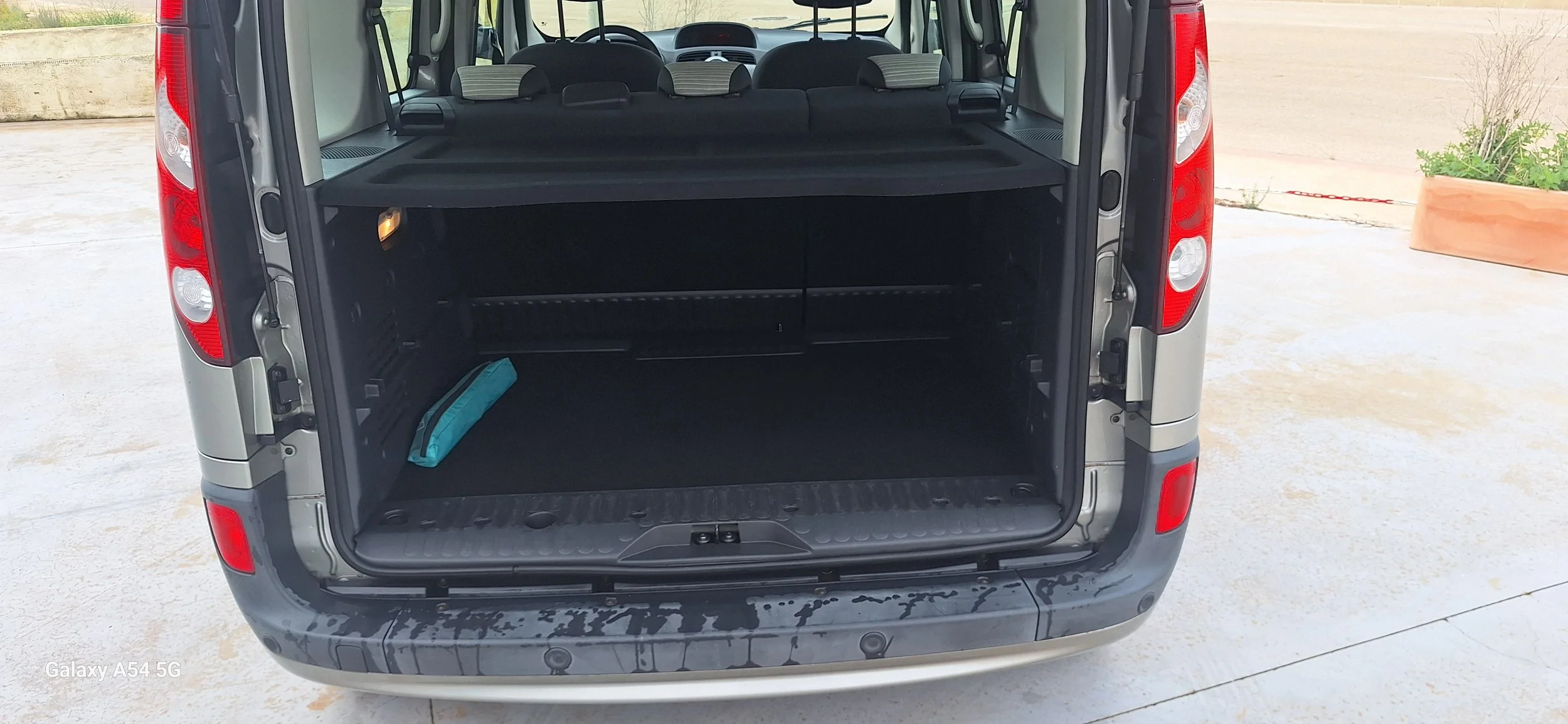 The open rear of a minivan showing the cargo area with a blue item on the left side and the back seats in an upright position. The minivan is parked on a concrete surface with a beige wall and a large planter with green plants in the background.