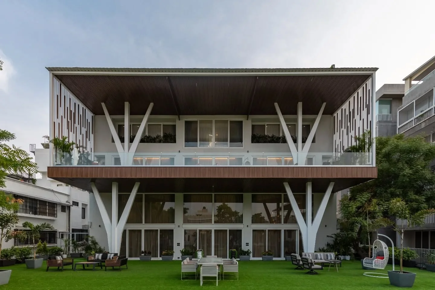 A white canvas, brushed with balance. From the white fa&ccedil;ade with its tree-branch columns to lawns layered across levels, this home doesn&rsquo;t hide its scale &mdash; it owns it. White walls, wood cladding, and layered lawns soften the edges,