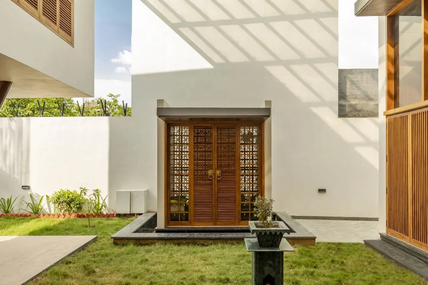 The courtyard is where the house reveals its core. A temple set within wood, jaali screens that let air pass freely, and a facade that opens rather than encloses. V-shaped columns add a certain sharpness, holding the space together while keeping it l
