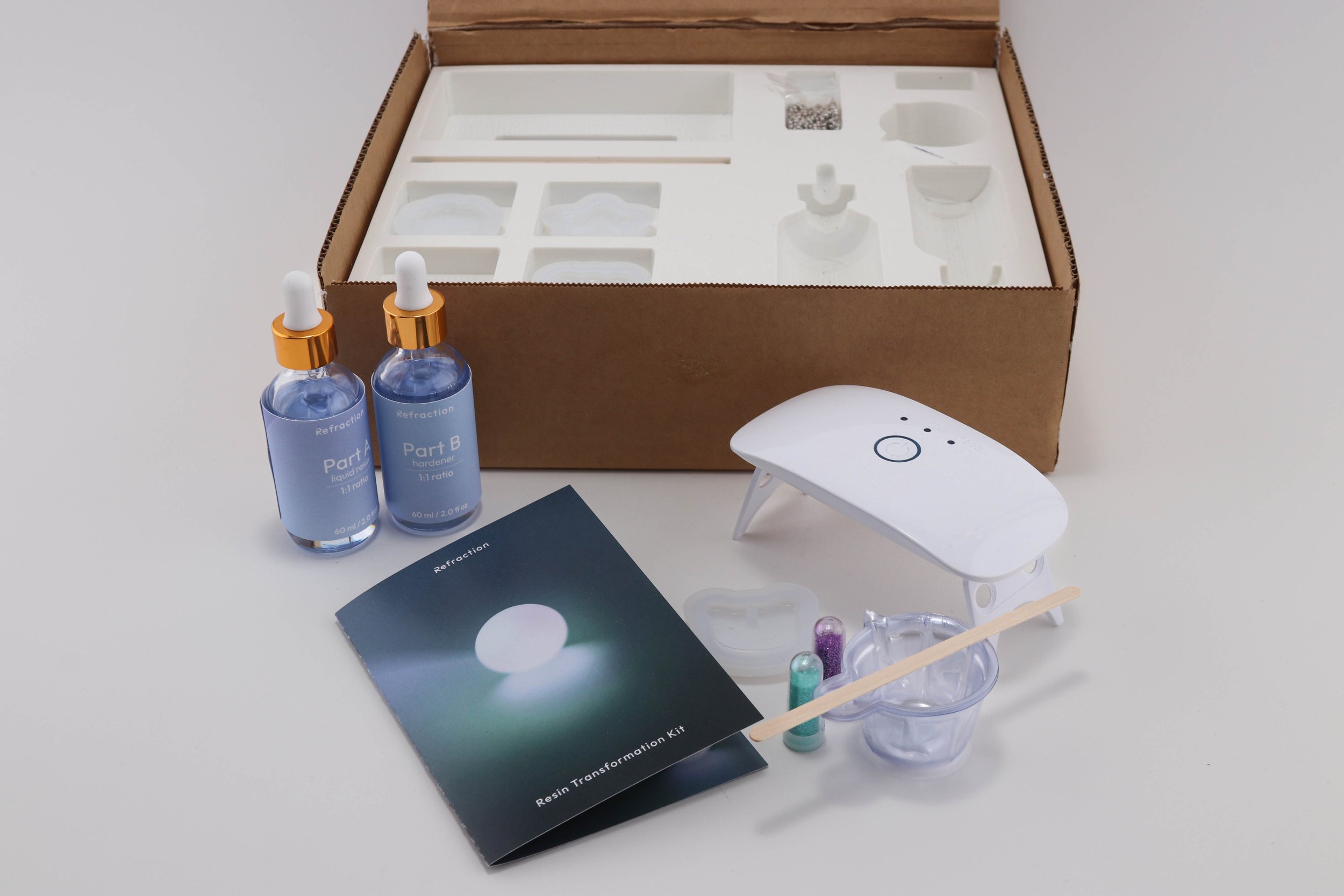 A resin transformation kit includes two bottles labeled 'Part A' and 'Part B', a pamphlet titled 'Resin Transformation Kit', a small white stool, a plastic cup with capsules of colored resin, a glass container, and a small beaker with purple liquid, all arranged on a white surface with a cardboard box and foam insert in the background.