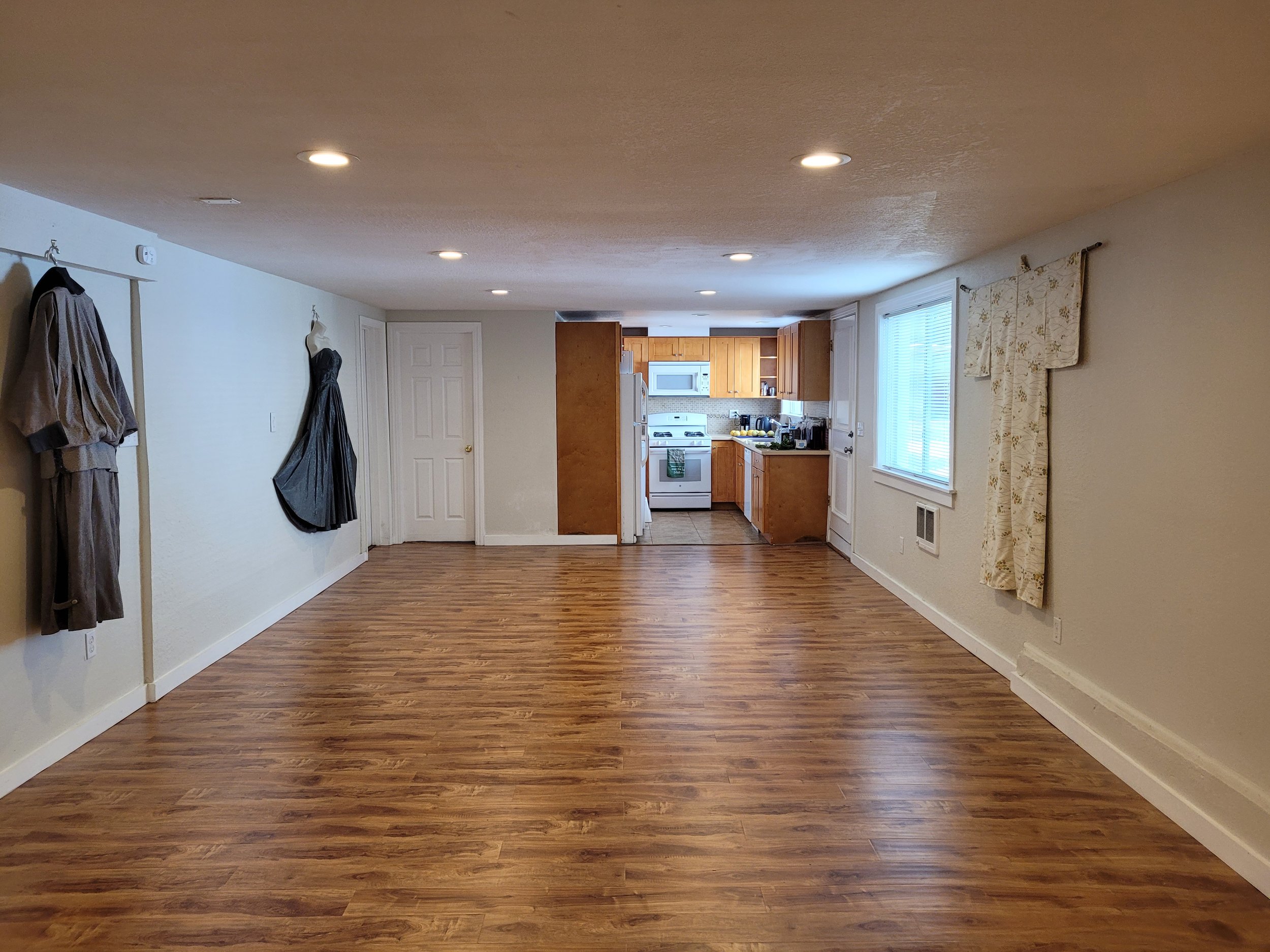 Room with wooden floor, coats hanging on the walls, kitchen with wooden cabinets in the background.