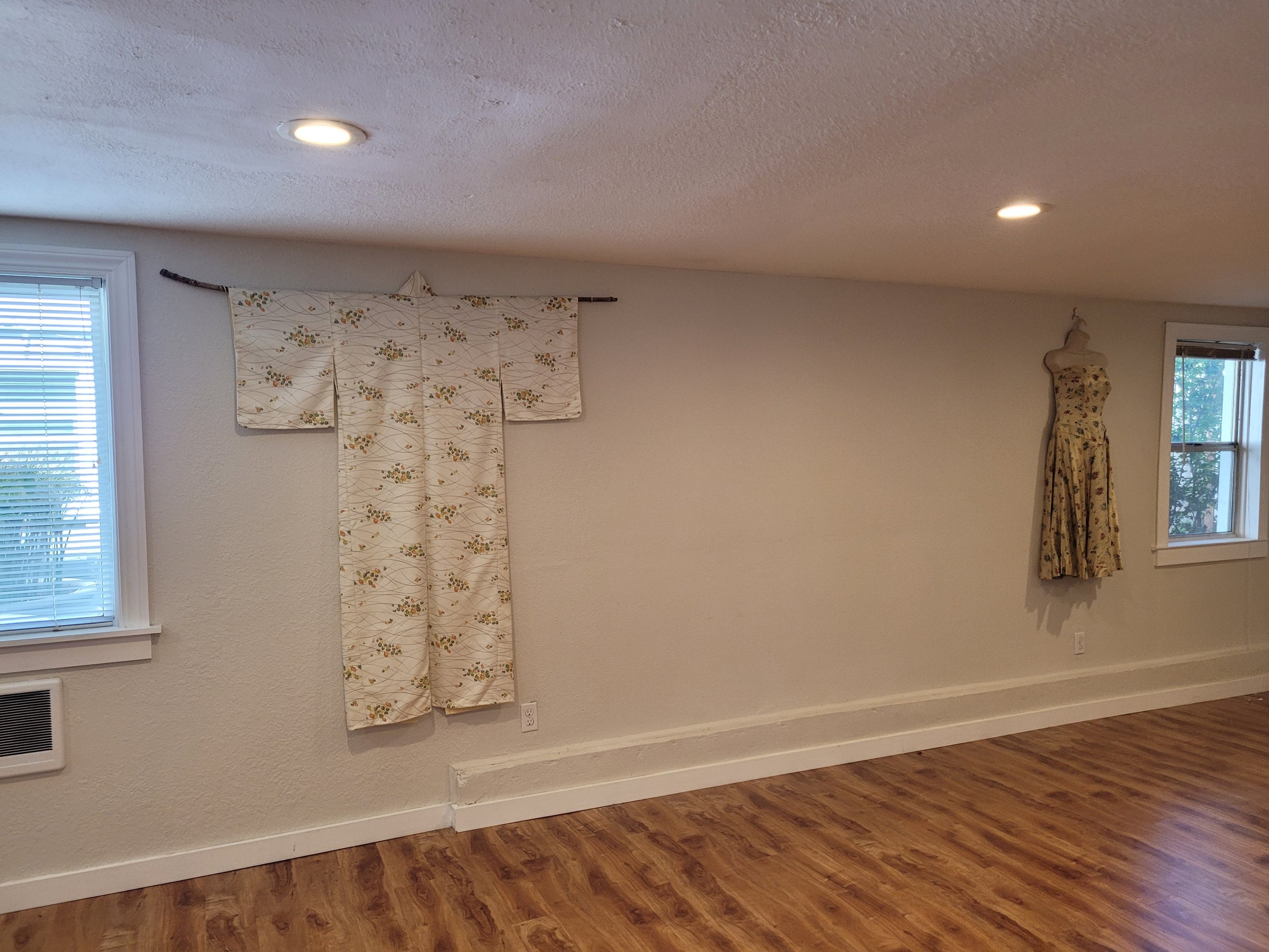Room with floral kimono on wall and dress on mannequin, wooden floor, windows, recessed lighting.