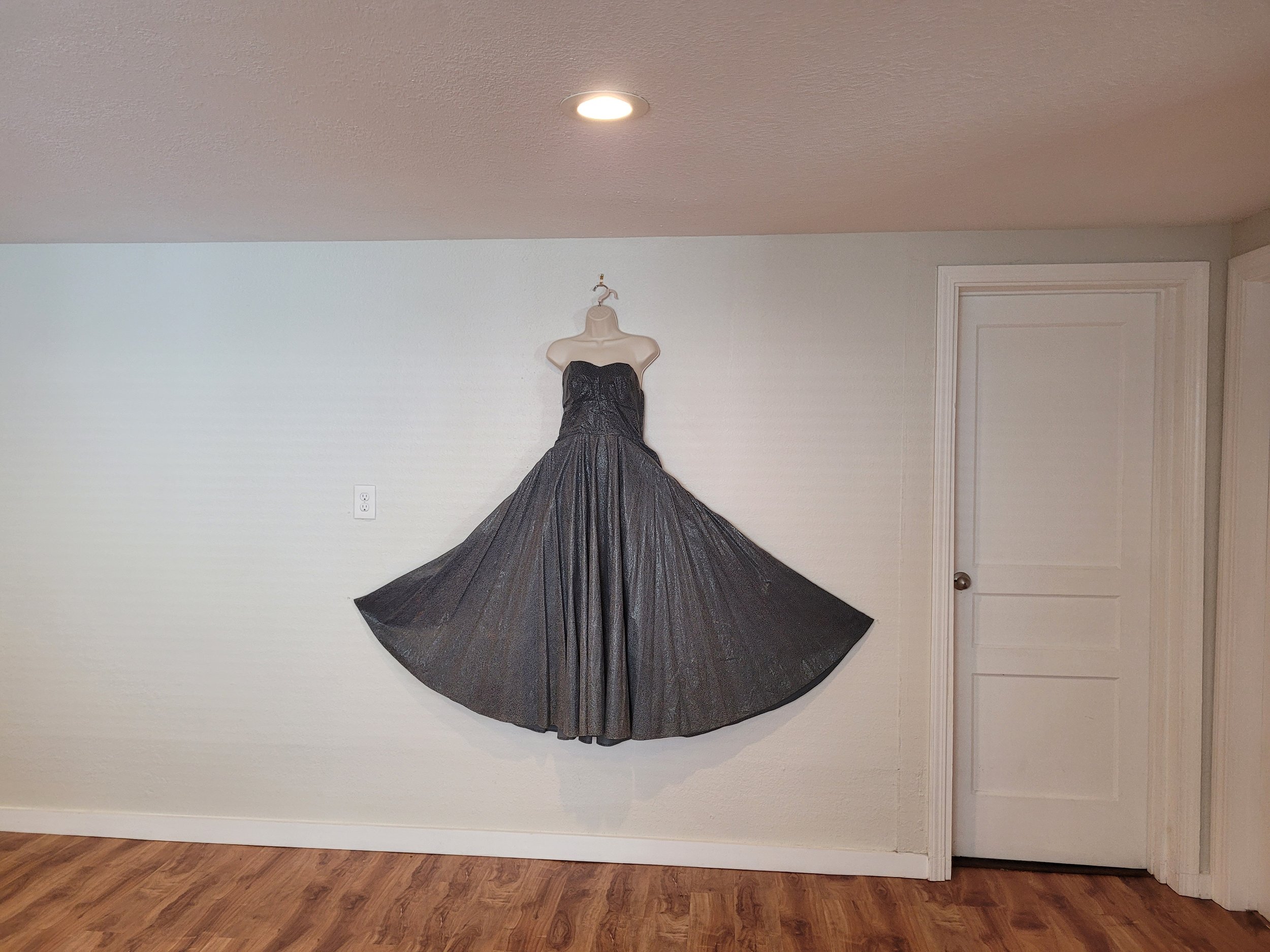 A strapless black dress displayed on a wall-mounted mannequin torso in a room with wooden flooring and white walls.