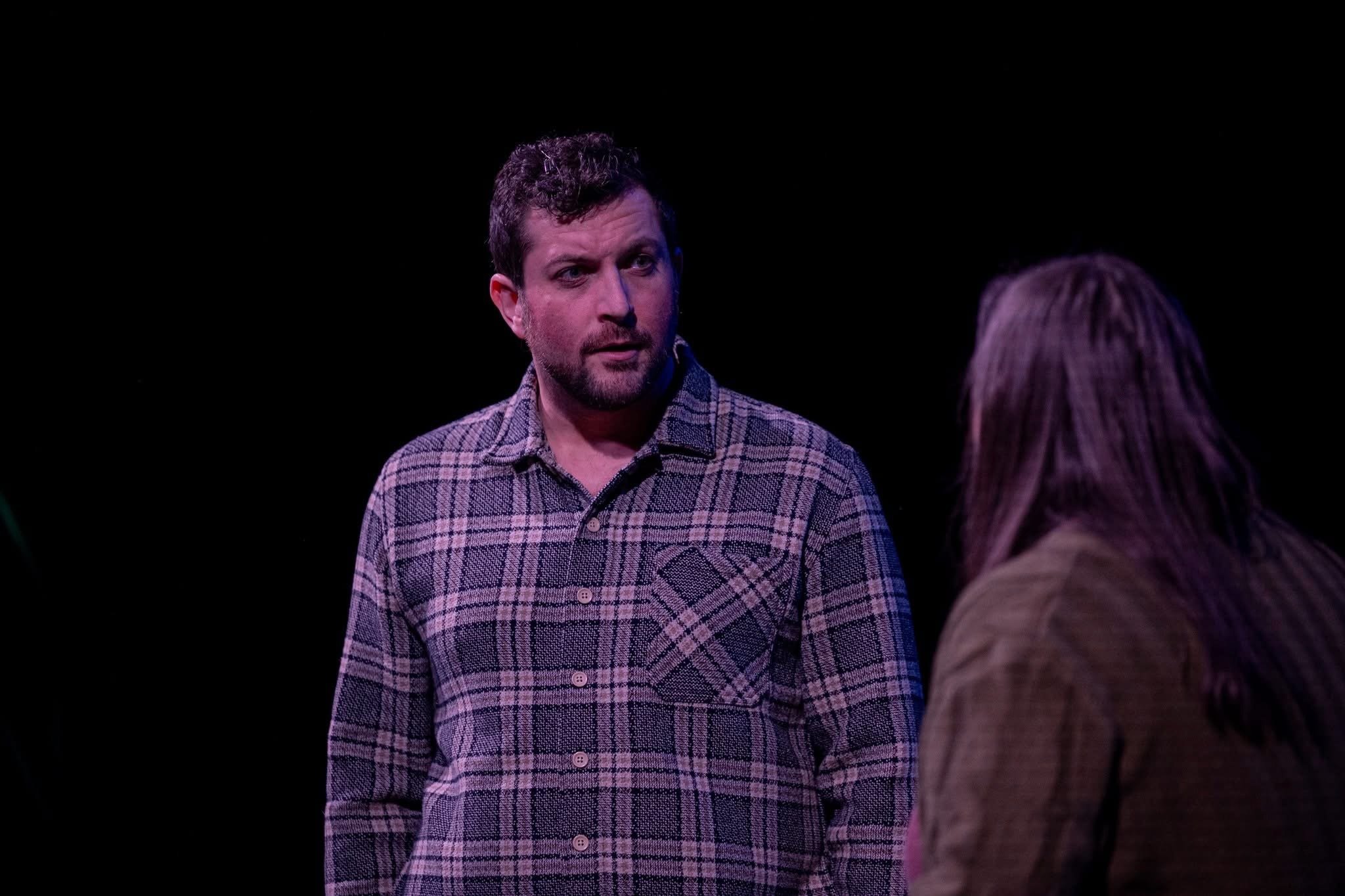 Two men engaged in a conversation on a dark stage. One man is facing the camera, wearing a plaid shirt, with a serious expression. The other man has his back to the camera, with long hair and a brown jacket.