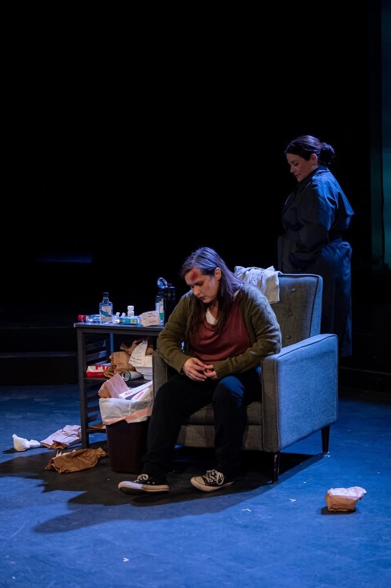 Two women on a stage, one sitting in a chair appearing upset, the other standing behind her with a somber expression. There is a table with trash and medicine bottles next to the chair.