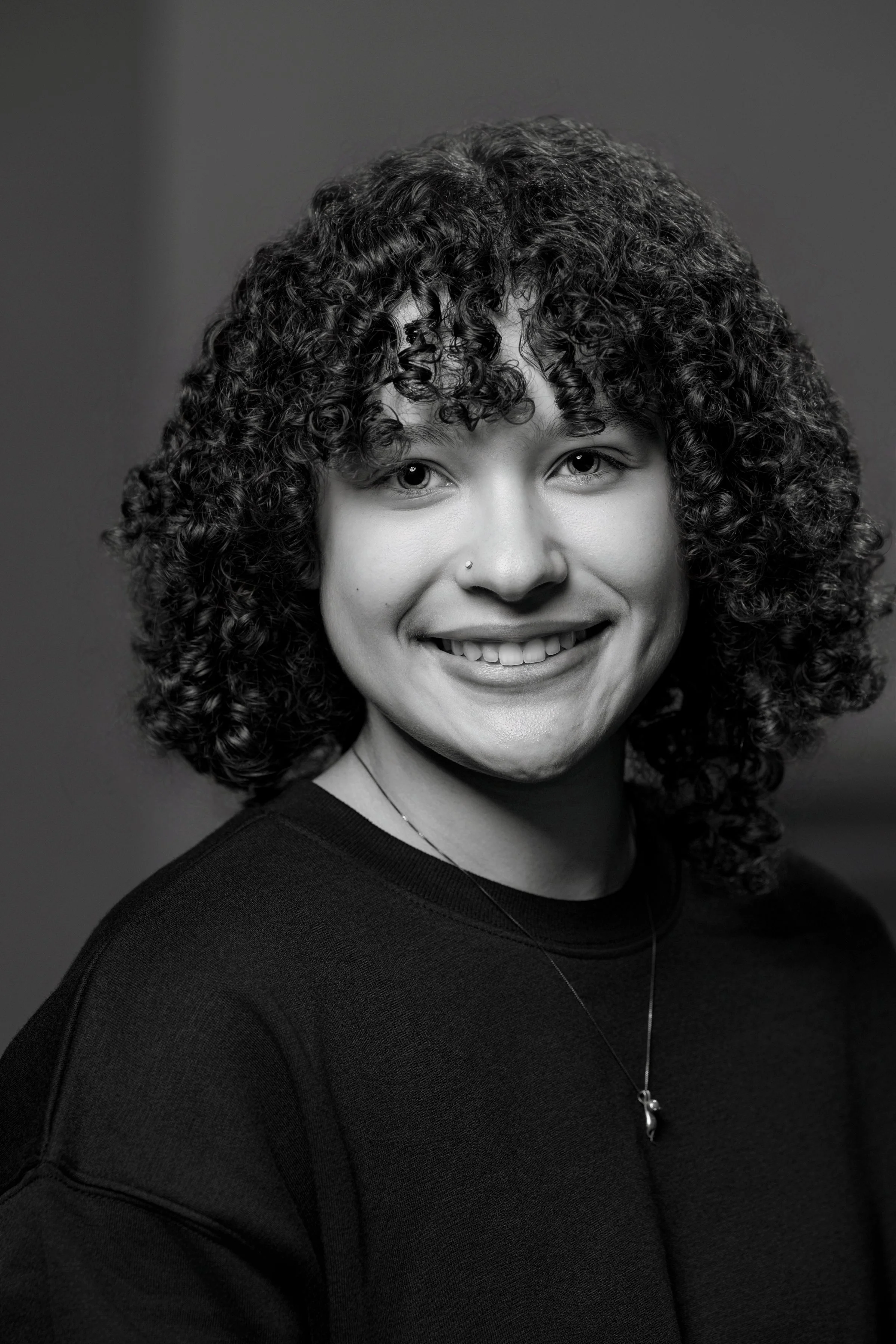 Black and white portrait of a woman with curly hair, smiling, wearing a dark top and a necklace with a small pendant.