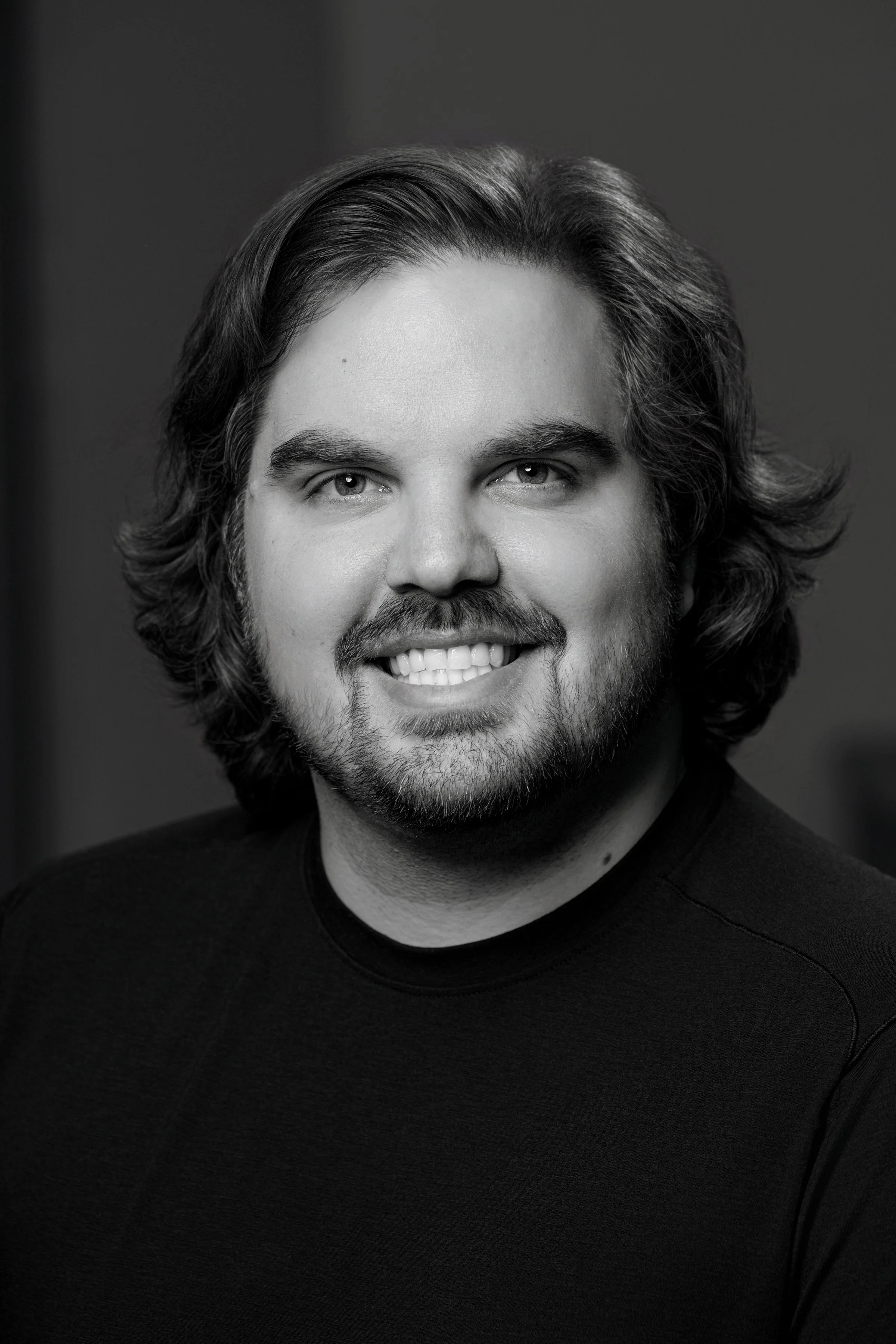Black and white portrait of a smiling man with wavy hair, beard, and short mustache, wearing a dark shirt.