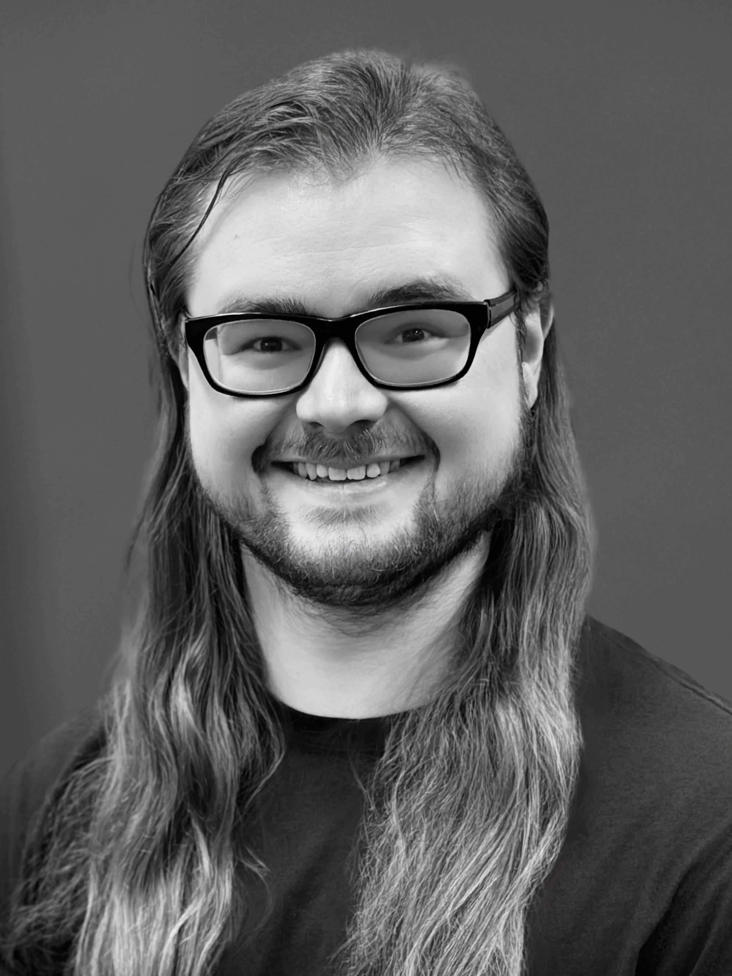 Black-and-white portrait of a smiling man with long hair, beard, mustache, glasses, wearing a dark shirt, against a plain background.