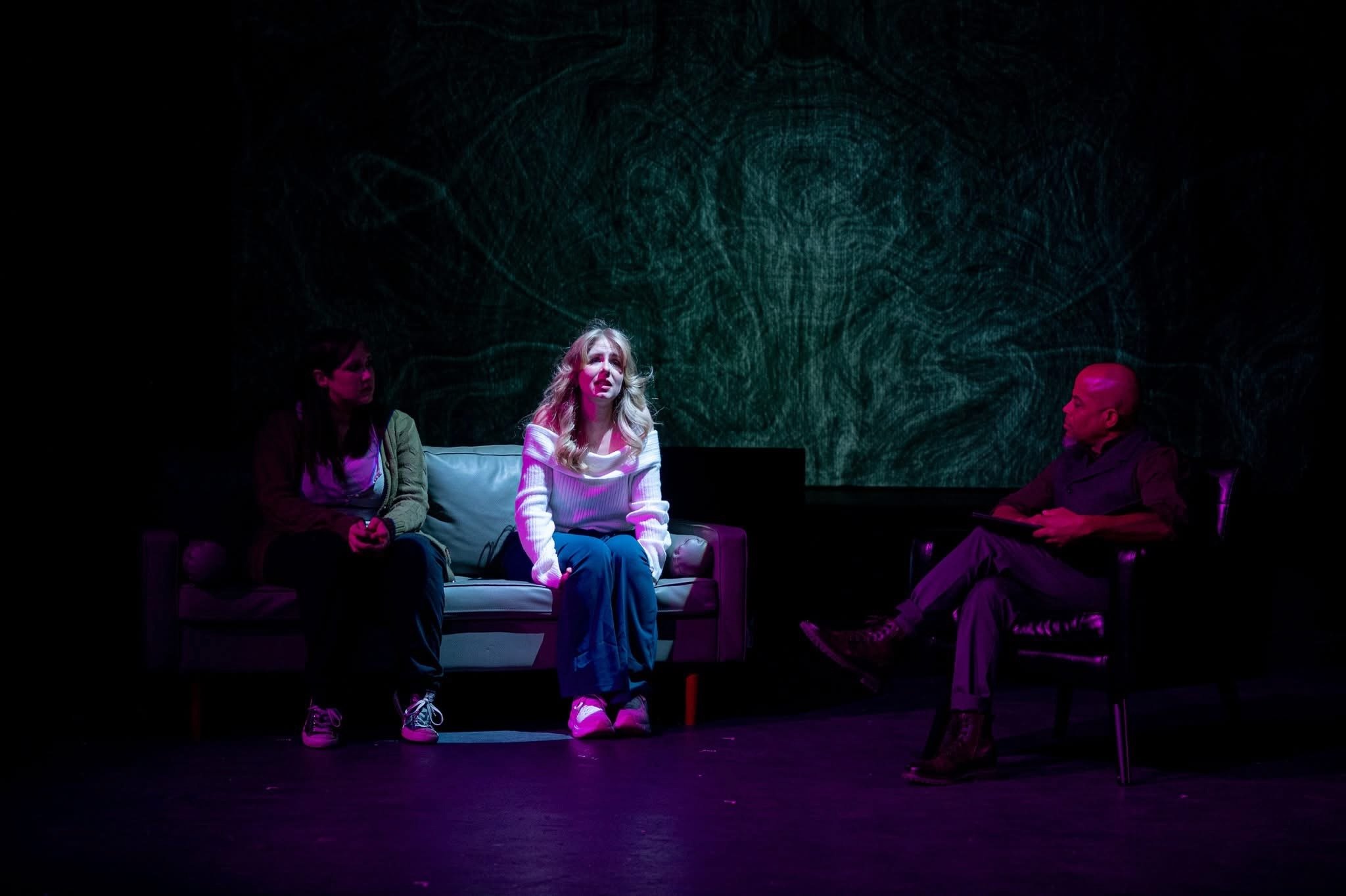 Three people are sitting on stage. Two women sit on a couch on the left, and a bald man sits on a chair on the right. The woman in the middle is illuminated with pink light, and the background features a green abstract design.
