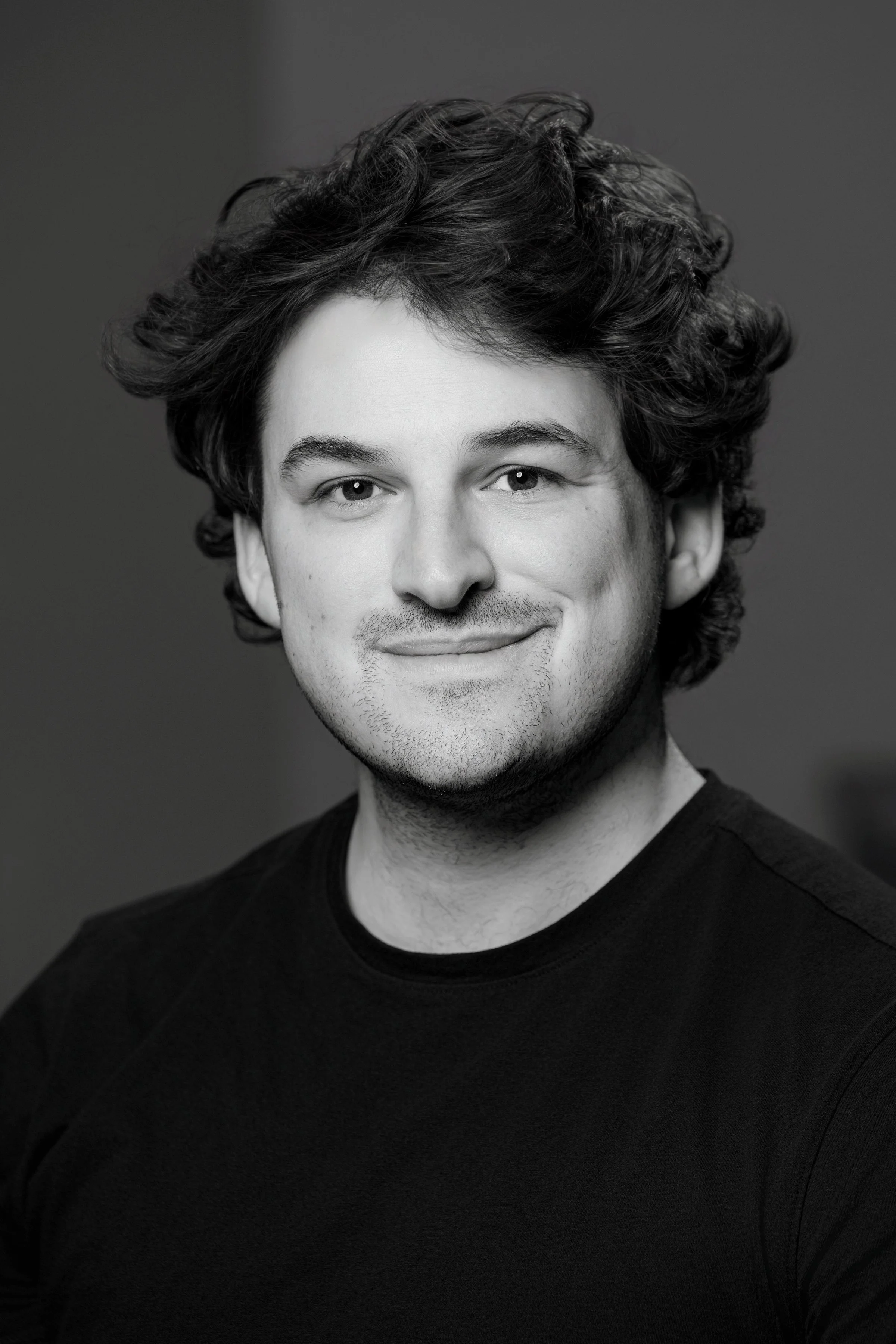 A black and white portrait of a young man with curly hair, wearing a dark shirt, smiling softly at the camera.