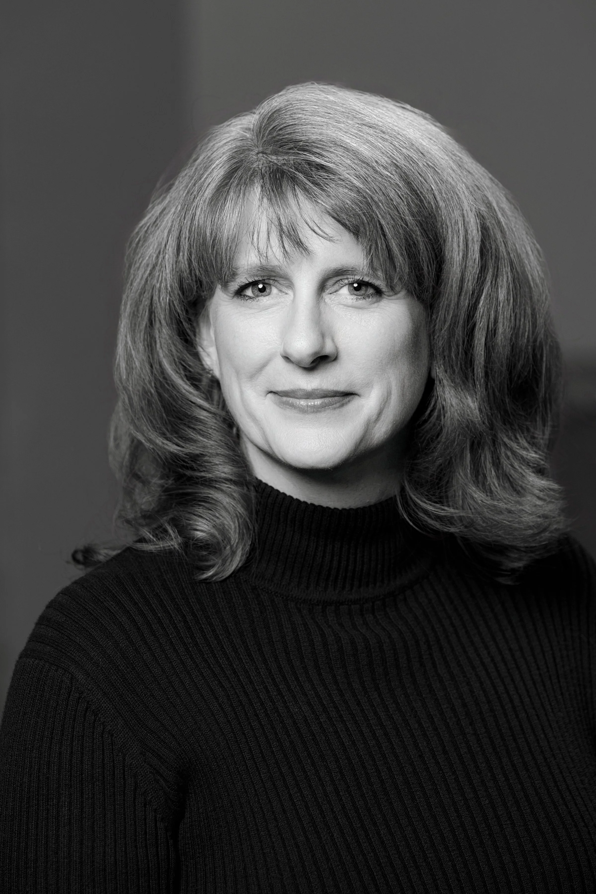A black-and-white portrait of a woman with shoulder-length wavy hair, wearing a ribbed turtleneck sweater, smiling slightly.