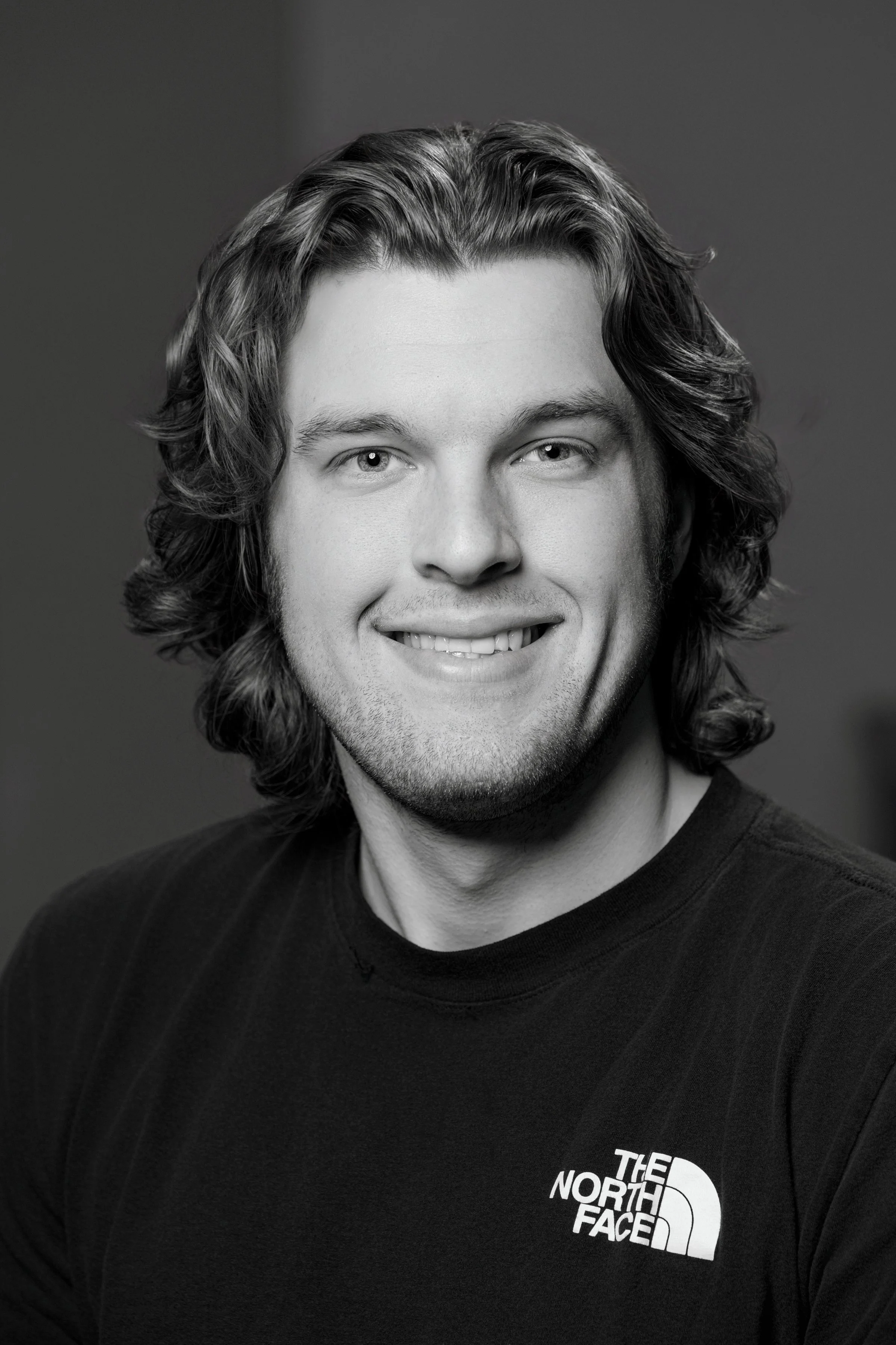 Black and white portrait of a young man with long wavy hair, smiling, wearing a T-shirt with The North Face logo.