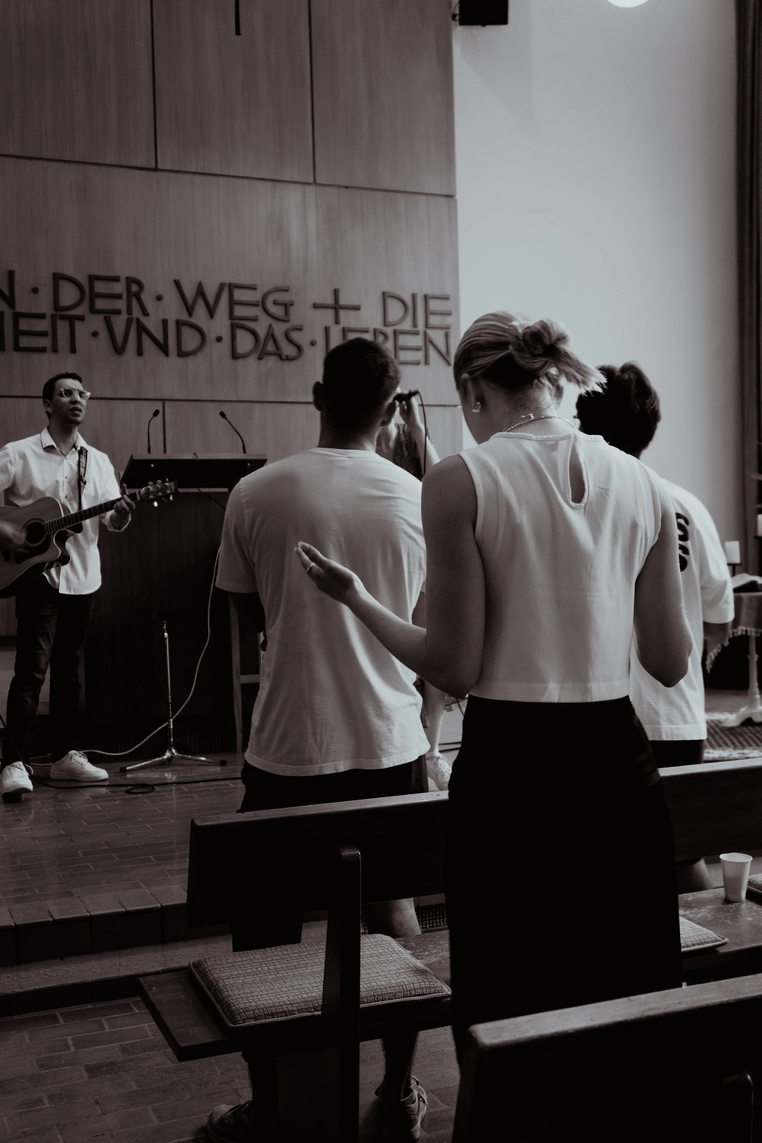 People praying or engaging in worship during a church service, with a man playing guitar and a woman singing on stage. Other attendees are standing and raising their hands in prayer.
