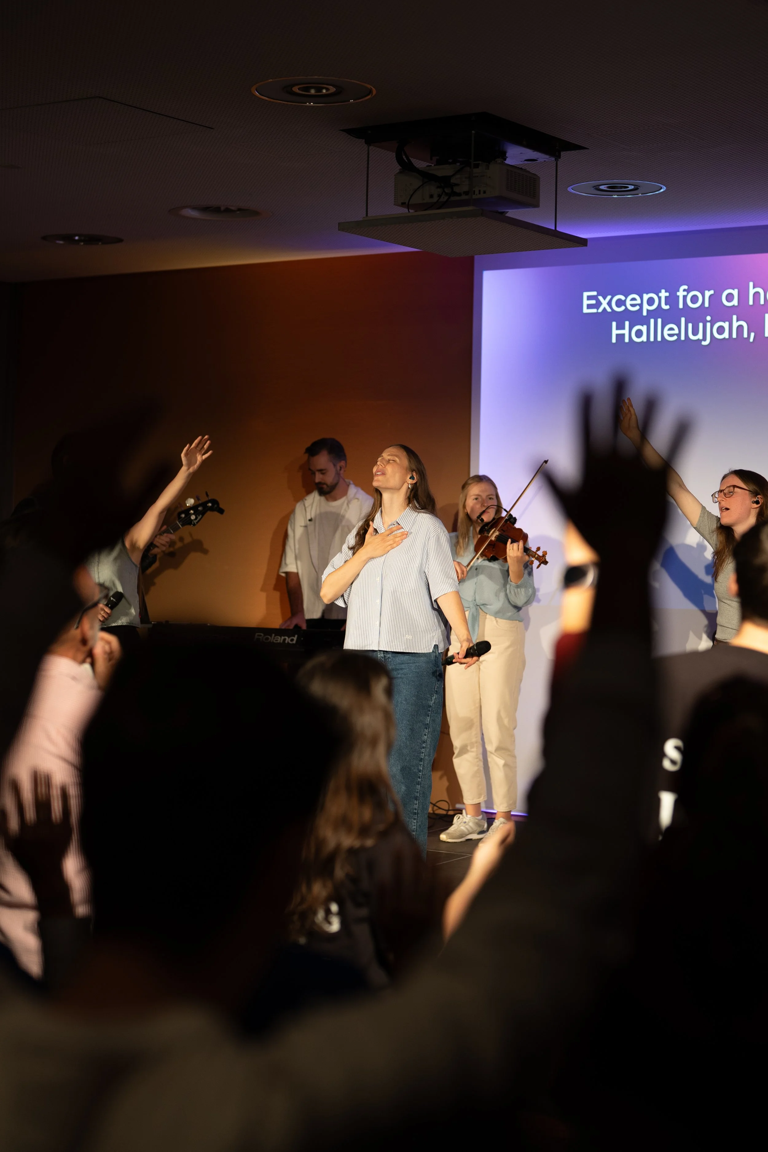 People praying or engaging in worship during a church service, with a man playing guitar and a woman singing on stage. Other attendees are standing and raising their hands in prayer.