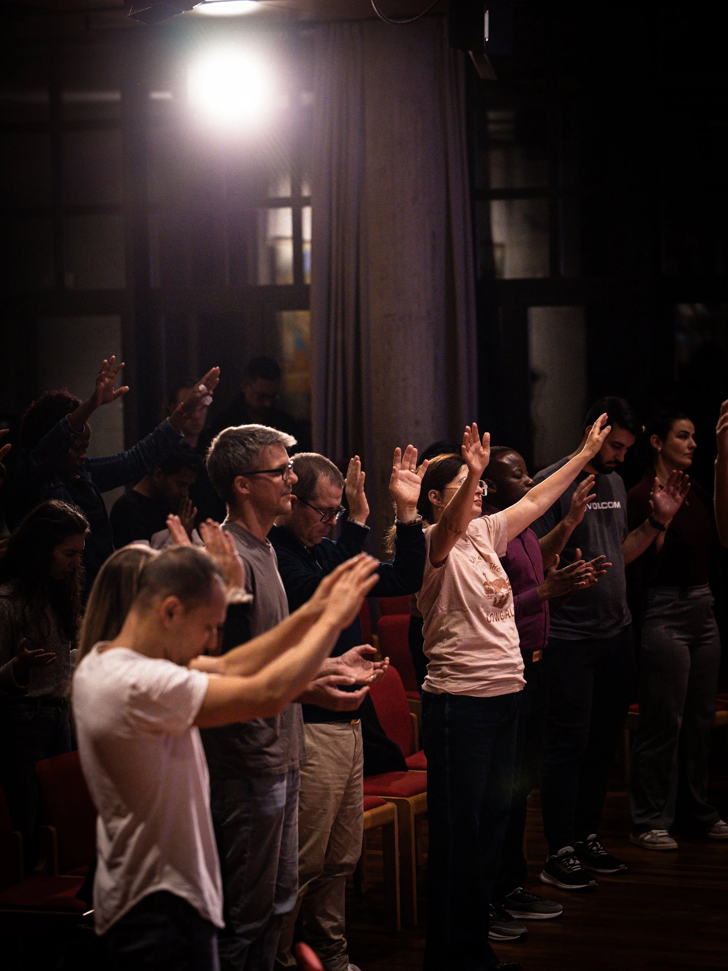 People standing with eyes closed and hands raised, participating in a worship or prayer service inside a dimly lit room with purple curtains and a bright light overhead.