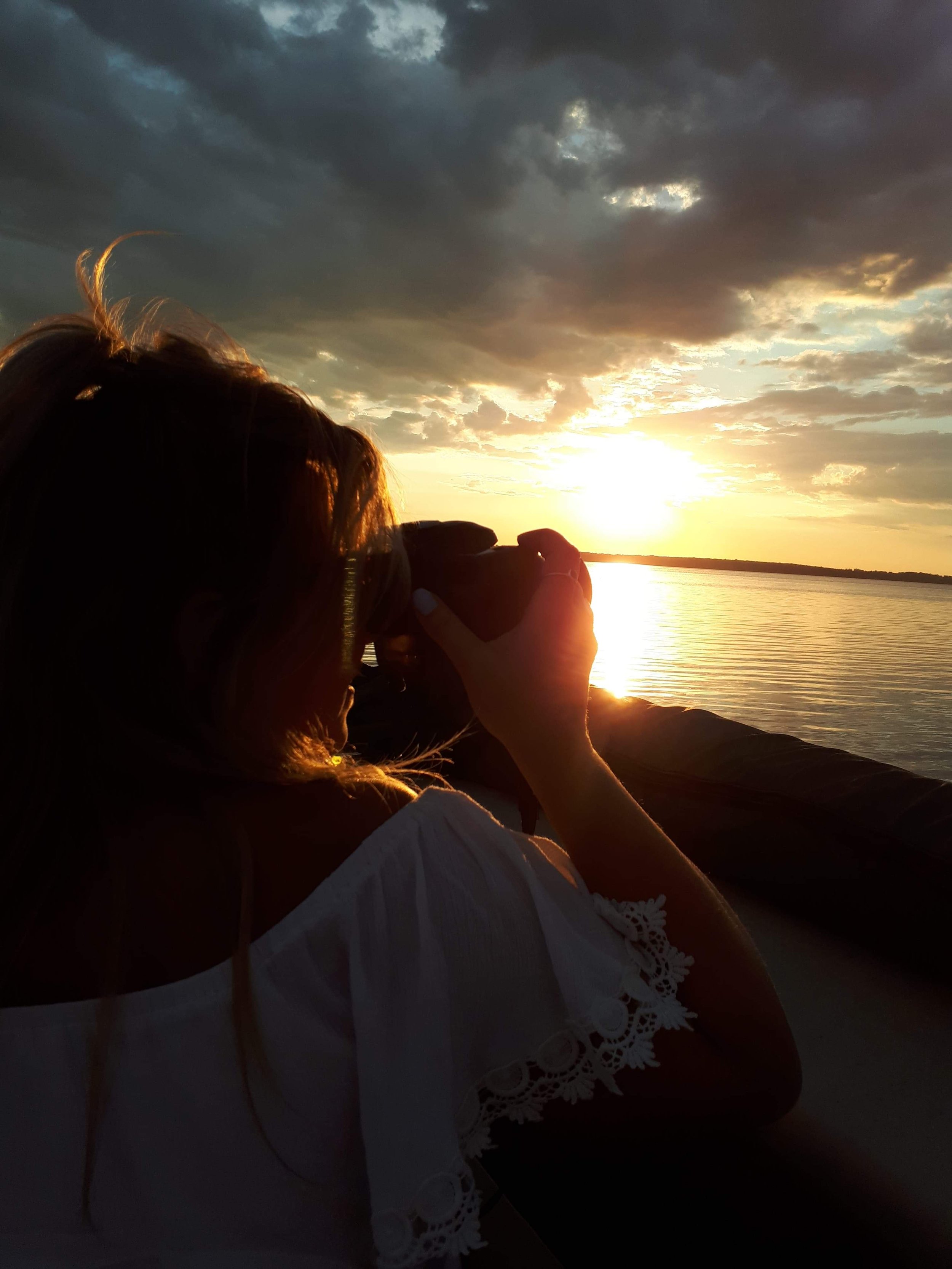 Person photographing a sunset over a body of water, with cloudy sky and orange hues, wearing a white off-shoulder dress.