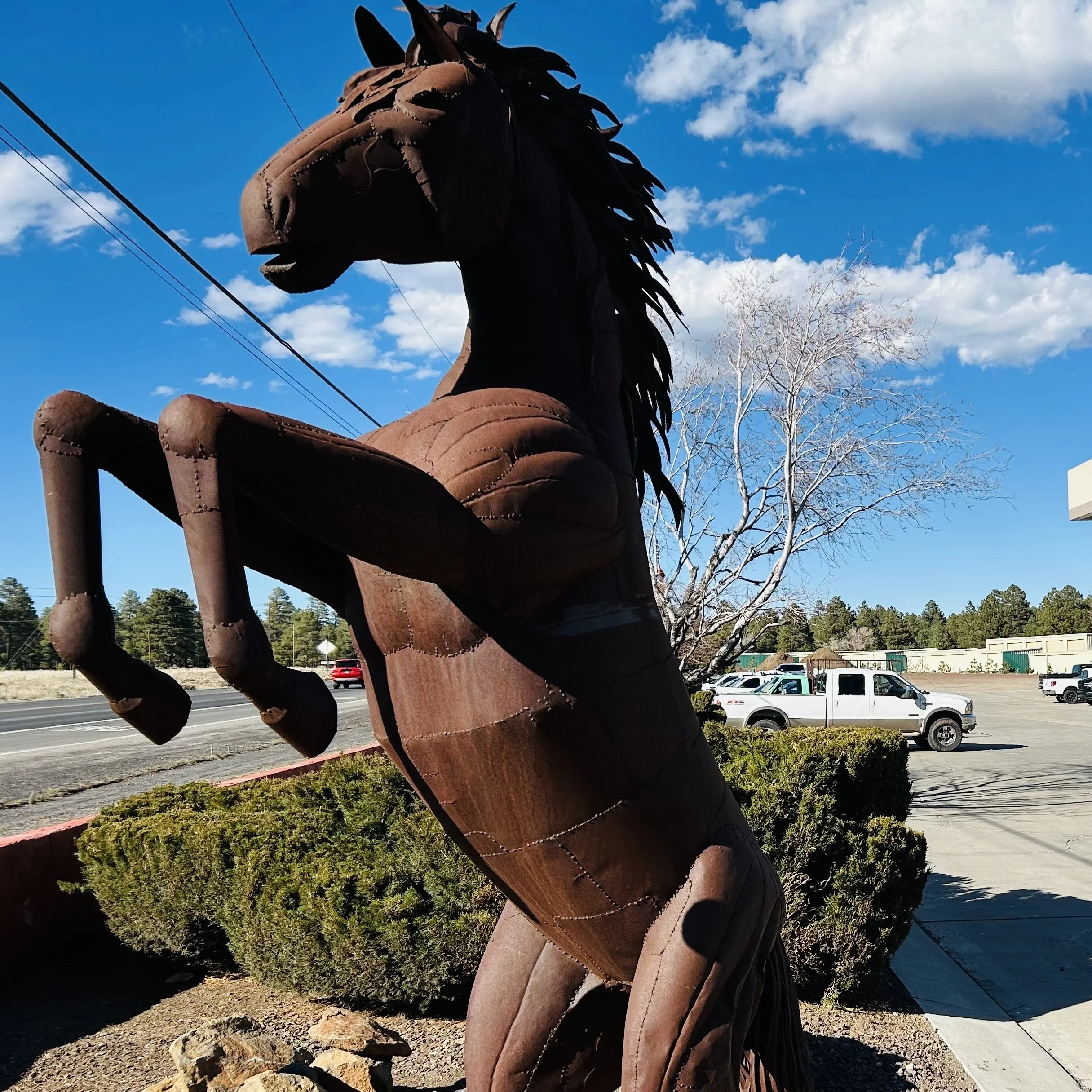 Large brown horse sculpture made of rod iron with dark mane and tail, standing outdoors near bushes, with trees, parked cars, and a blue sky with clouds in the background.