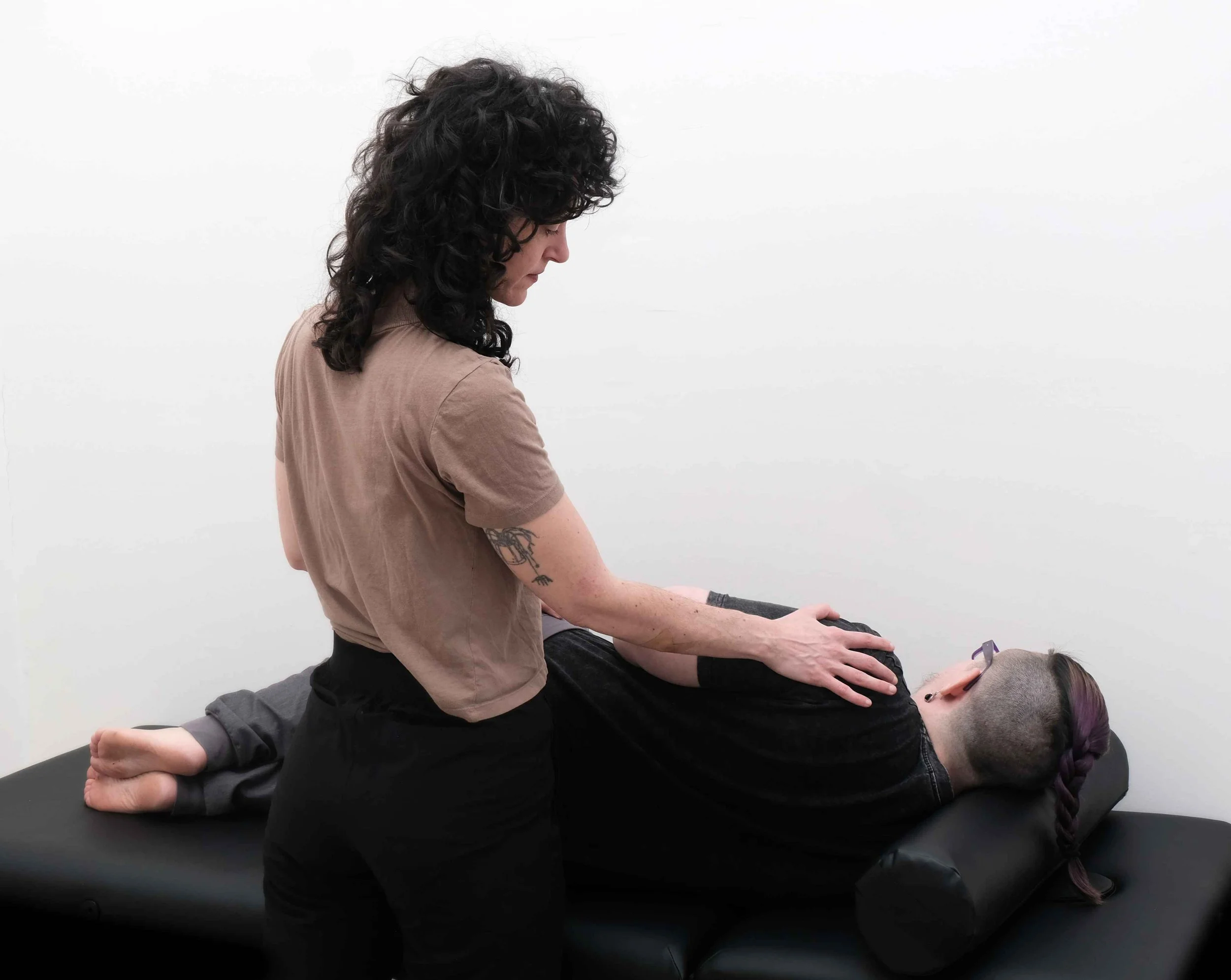 pelvic floor muscle lengthening in sidelying for chronic pelvic pain treatment, transmasc patient is lying on his right side, pelvic floor therapist is using their hands to give biofeedback to soften shoulders and neck to release neck tension