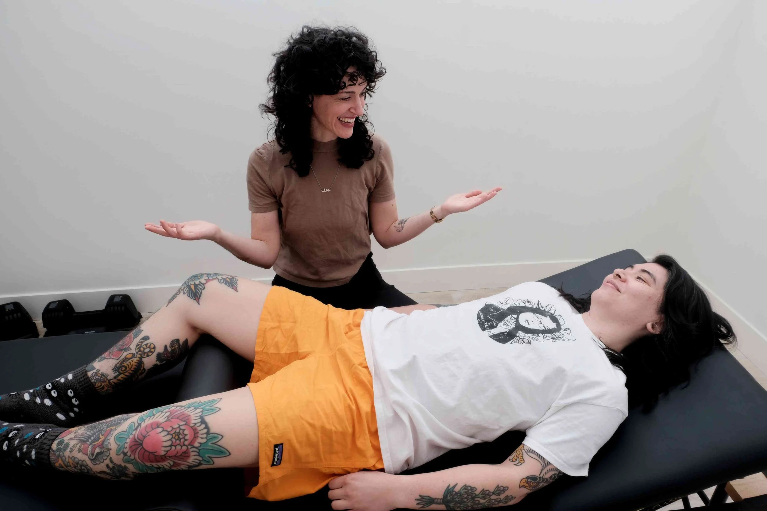 queer pelvic floor therapist with supine transmasculine qpoc patient, both smiling, discussing trauma informed healthcare for gender and sexual minorities.