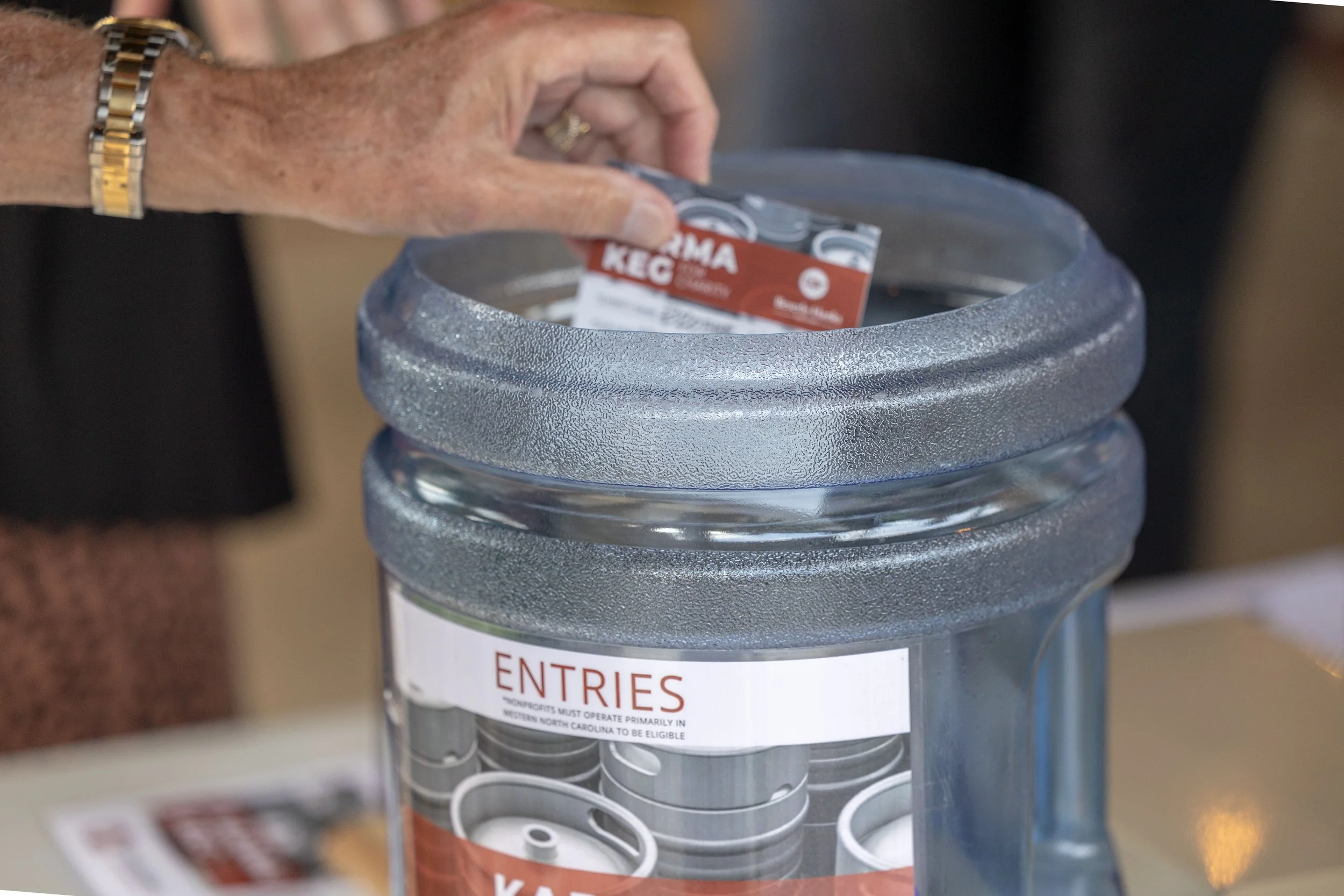 A hand dropping a K-4 Idaho Keg into a clear water cooler with a foam cover, marked with a sign that says 'ENTRIES'.