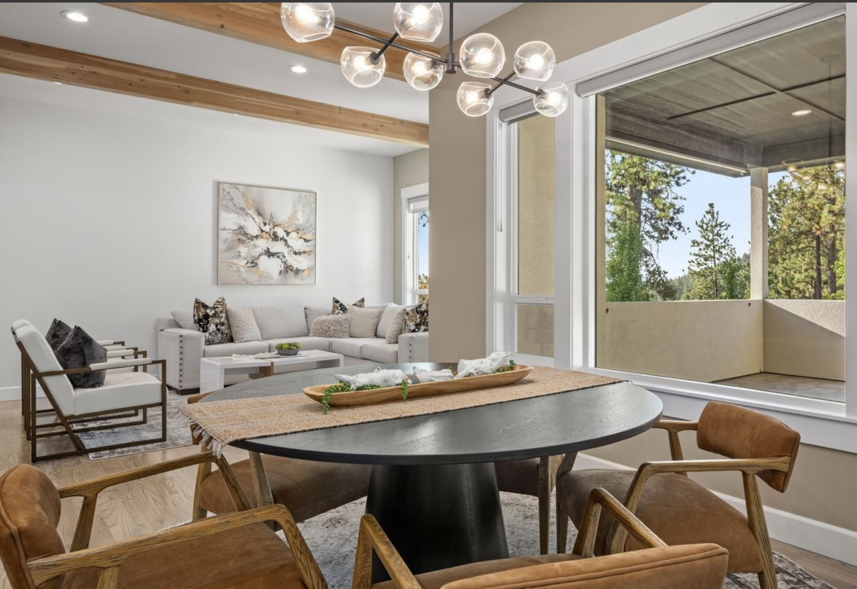 Professionally staged open concept dining and living area in the Inland Northwest, featuring a round black table with tan leather chairs, neutral sofa with pillows, exposed wood beams, abstract art and large windows overlooking pine trees