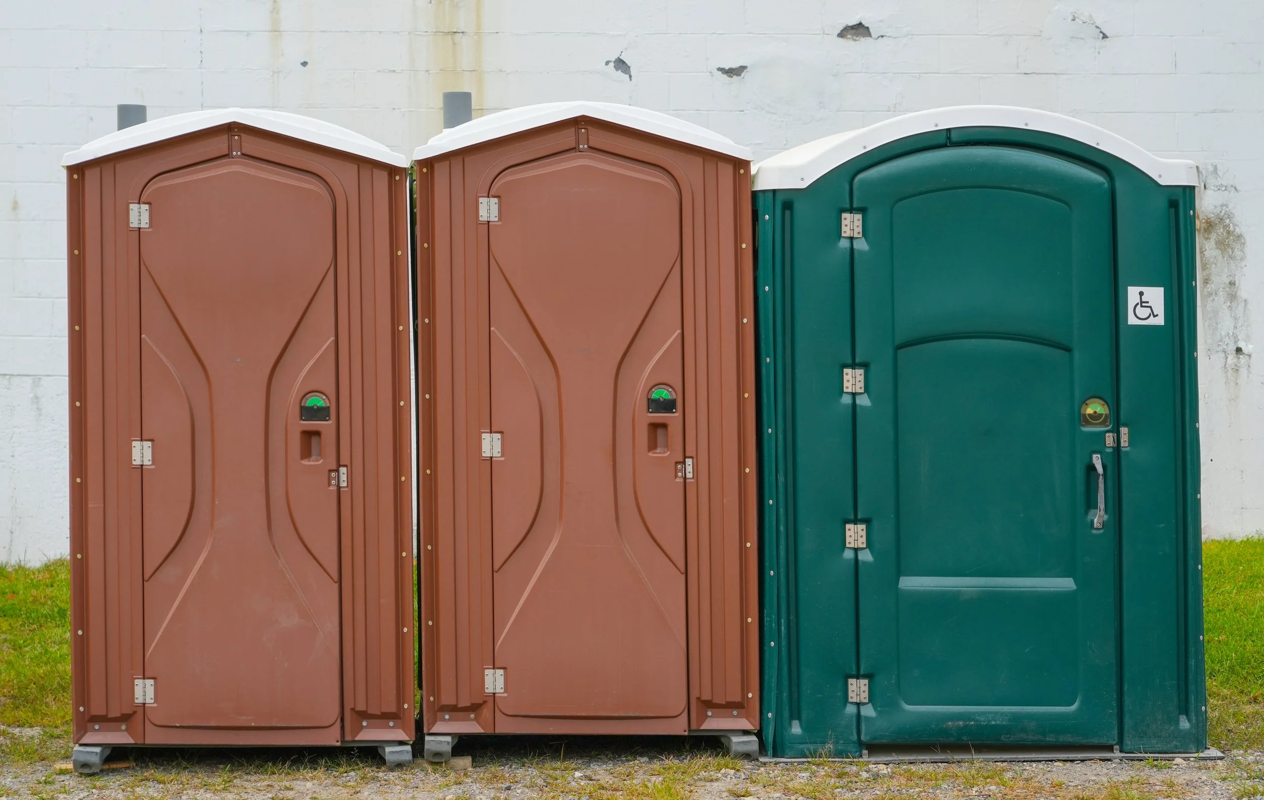 Three portable toilets side by side in front of a white brick wall, with grass and gravel in the foreground.