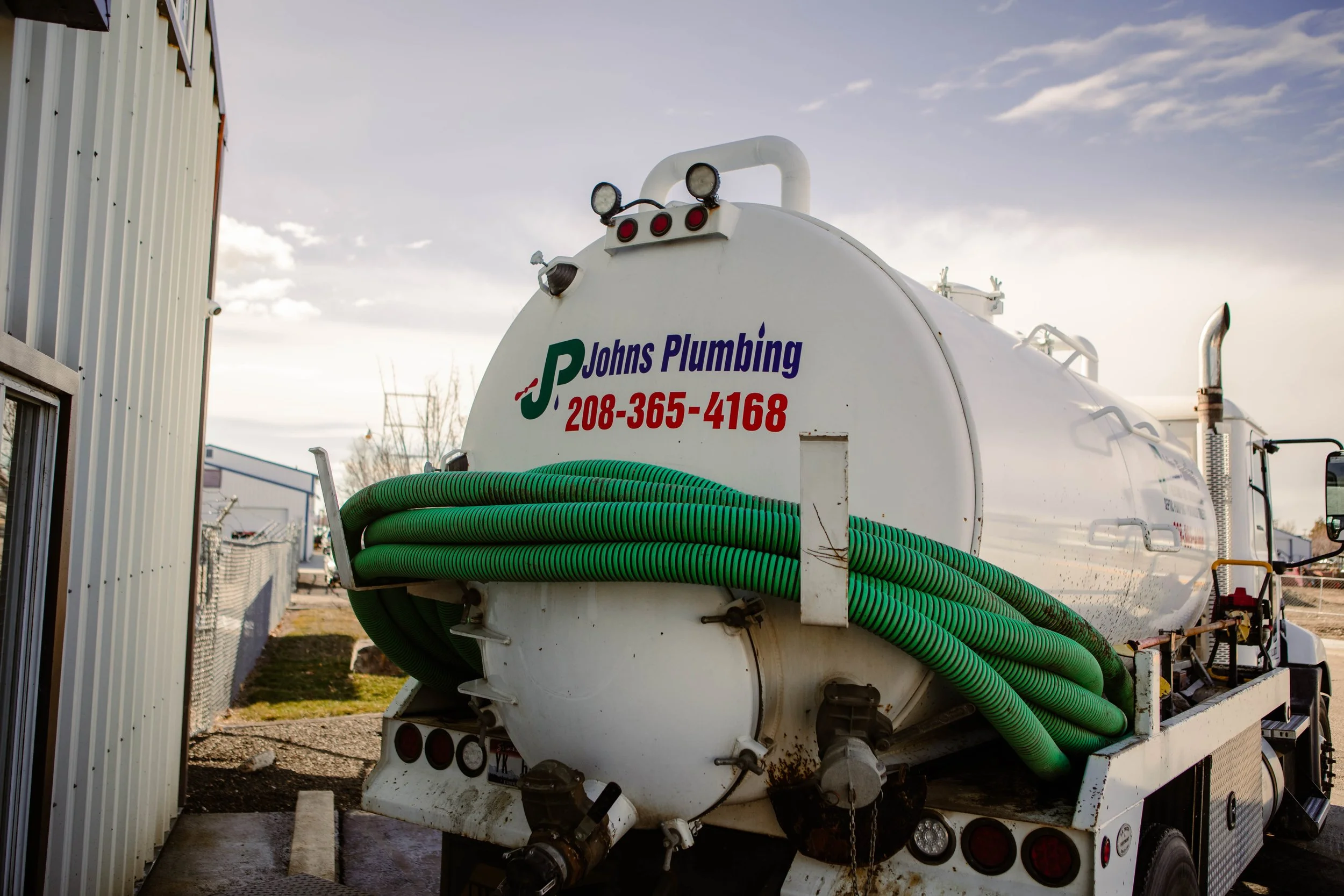 Back of a white plumbing truck with green hoses coiled on it, displaying contact info 'Johns Plumbing 208-365-4168'.