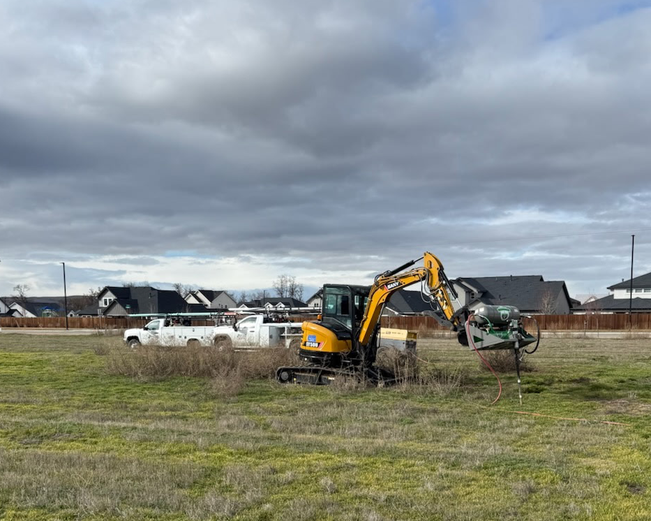 A small excavator operating on a grassy field, with an earth buster, under a cloudy sky, and residential houses in the background.