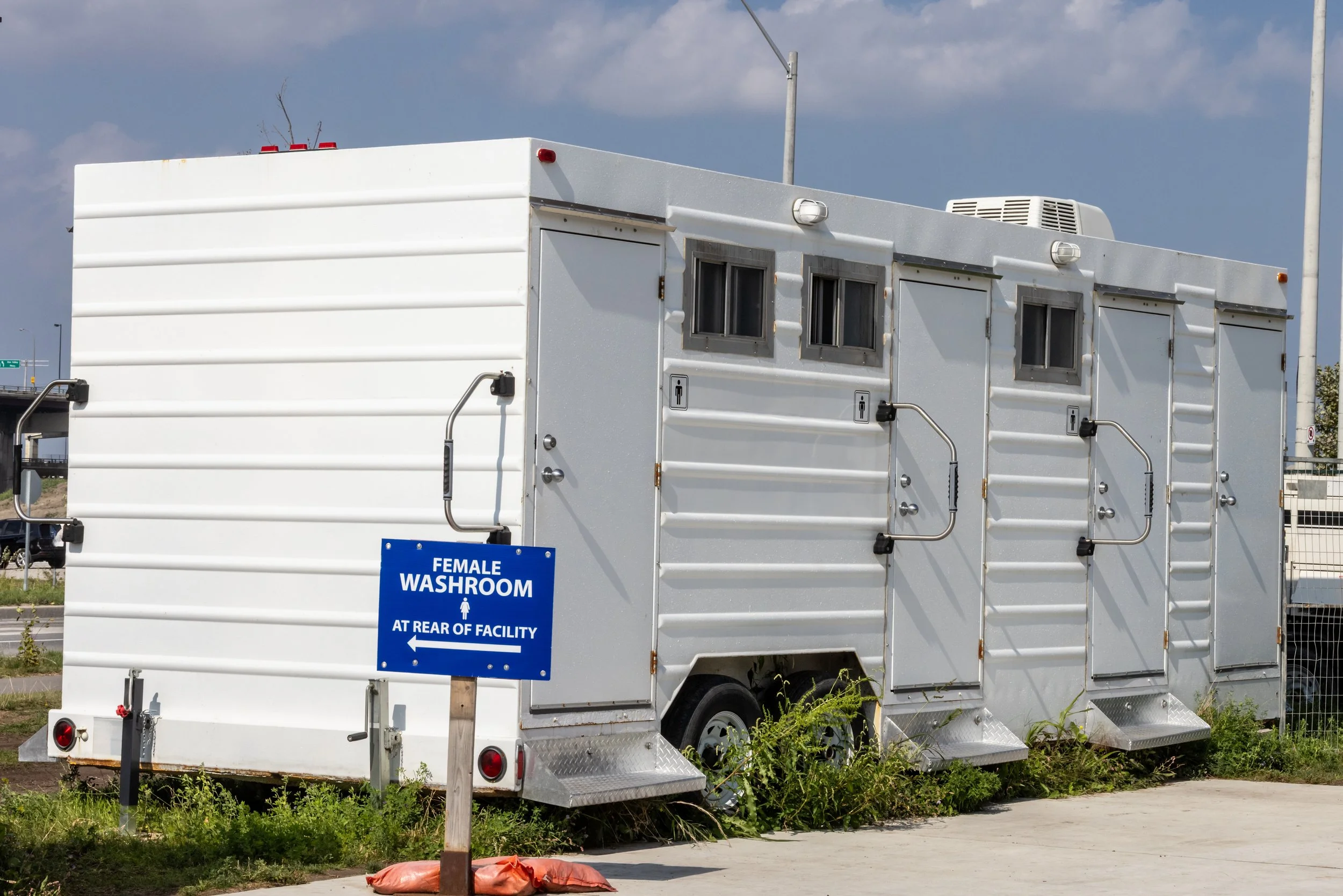 A white mobile restroom trailer with multiple doors and small windows, parked outdoors with a blue female washroom sign in front indicating it is at the rear of the facility.