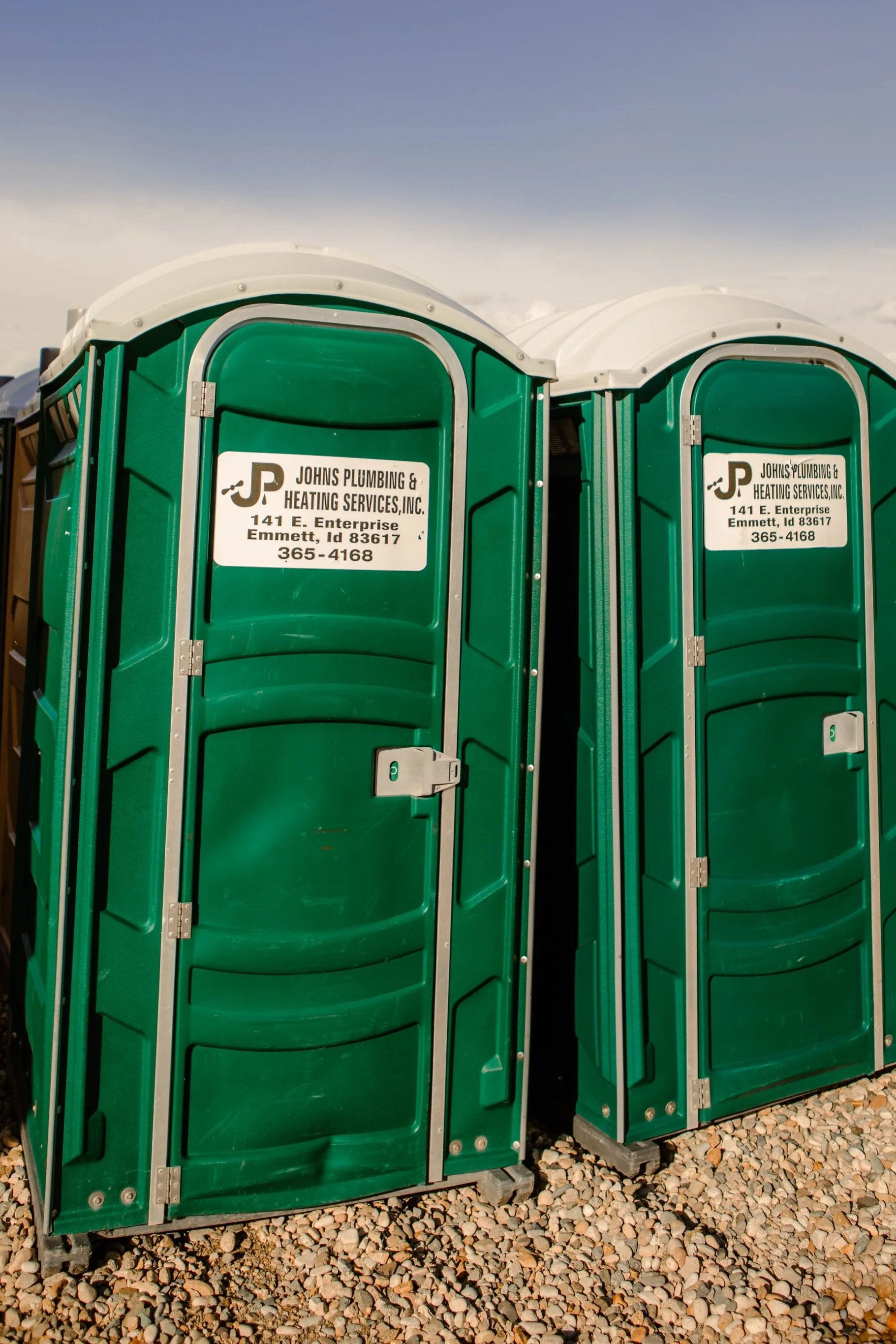 Two green portable toilets with white roofs, placed on a gravel surface, with a sign reading 'Johns Plumbing & Heating Services Inc., 141 E. Enterprise, Emmett, ID 83617, 365-4168'.