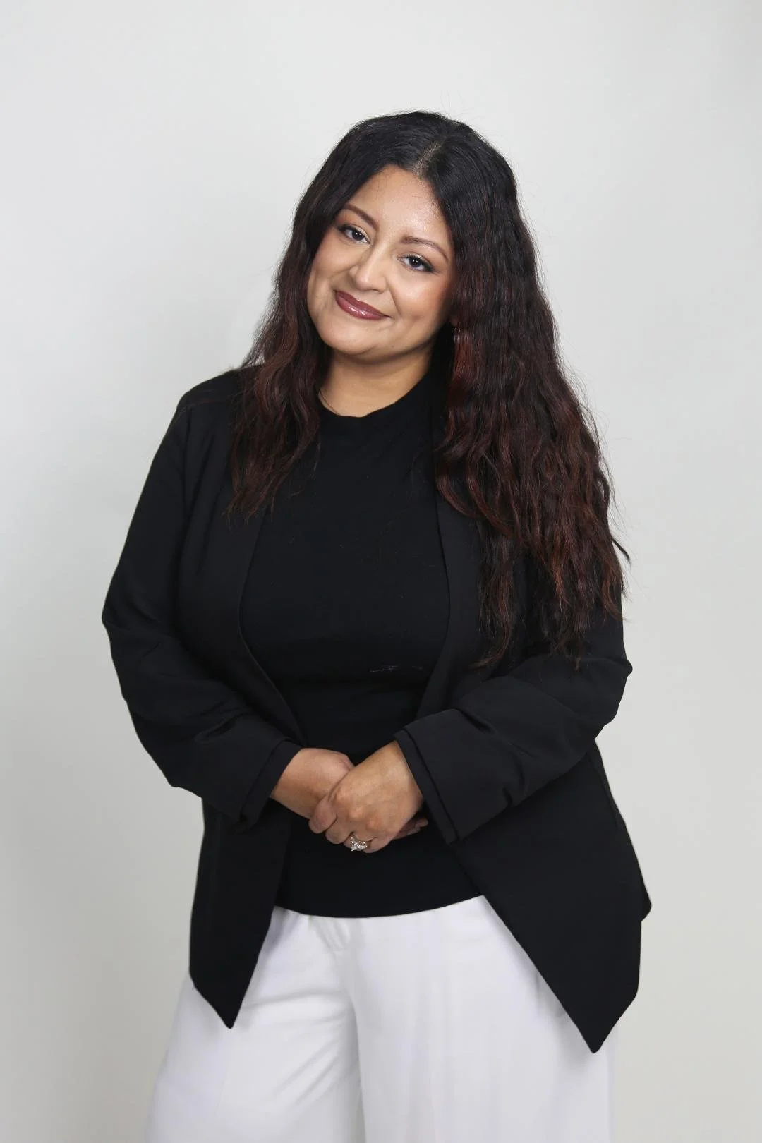 Portrait of a woman with long, dark hair wearing a black blazer and white pants against a neutral background.