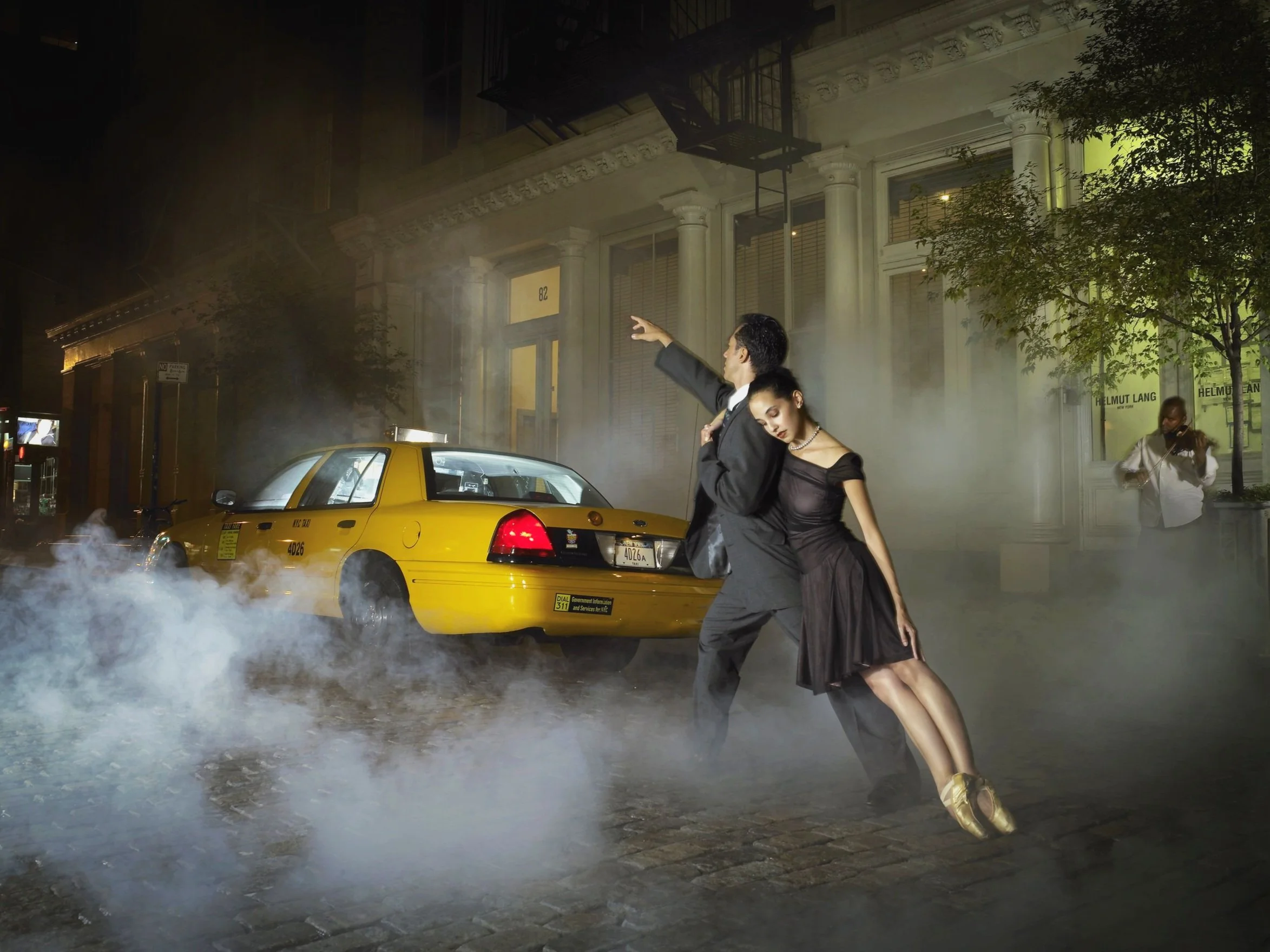 A man and woman dance ballet on a city street at night, with a yellow taxi and smoke in the background, and another person playing violin nearby.