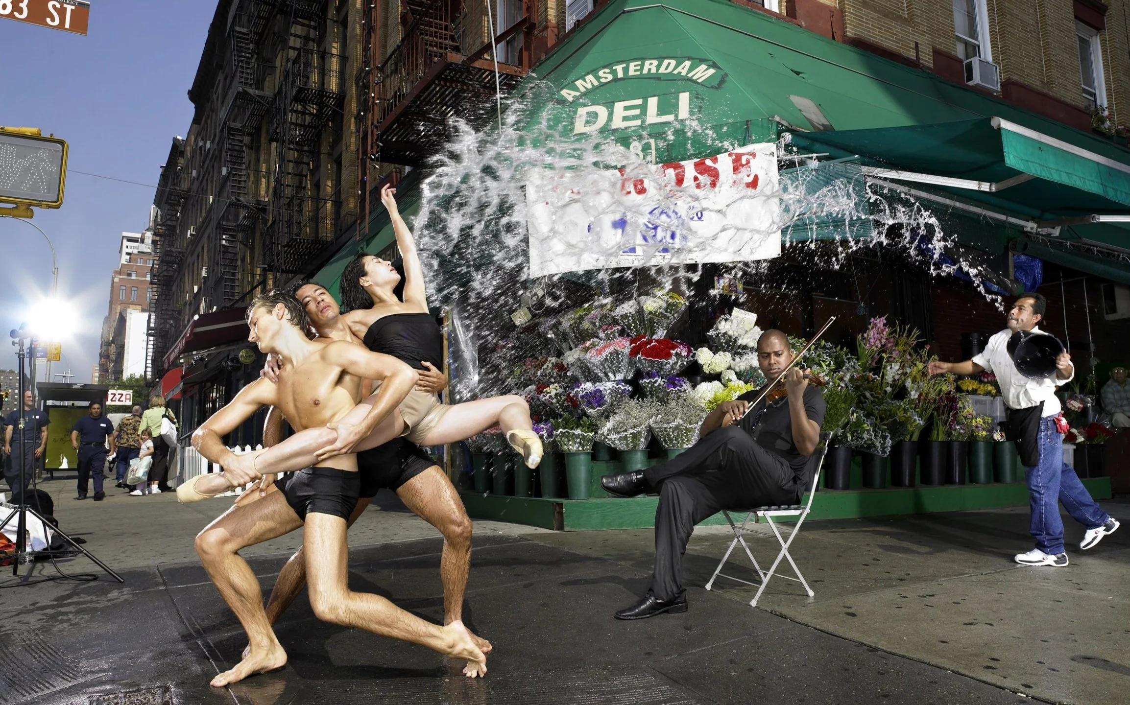 Ballet dancers perform on a city street with a flower shop in the background, while water is splashed onto them, and musicians play nearby.