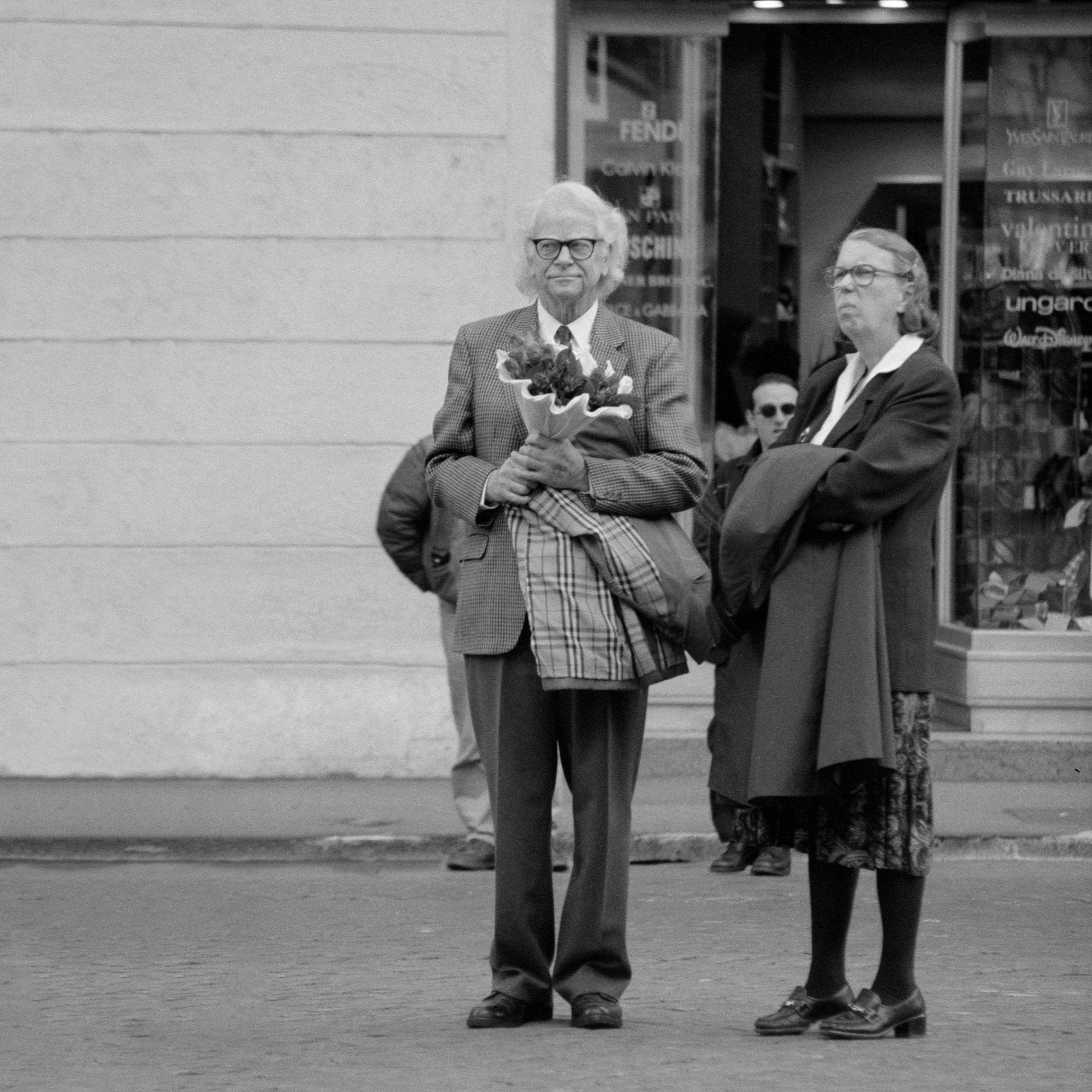 A black-and-white photograph of an older man holding a bouquet of flowers, standing on a sidewalk next to a woman with crossed arms. The man is wearing glasses, a plaid blazer, and dress pants, while the woman is dressed in a coat, skirt, and heels,