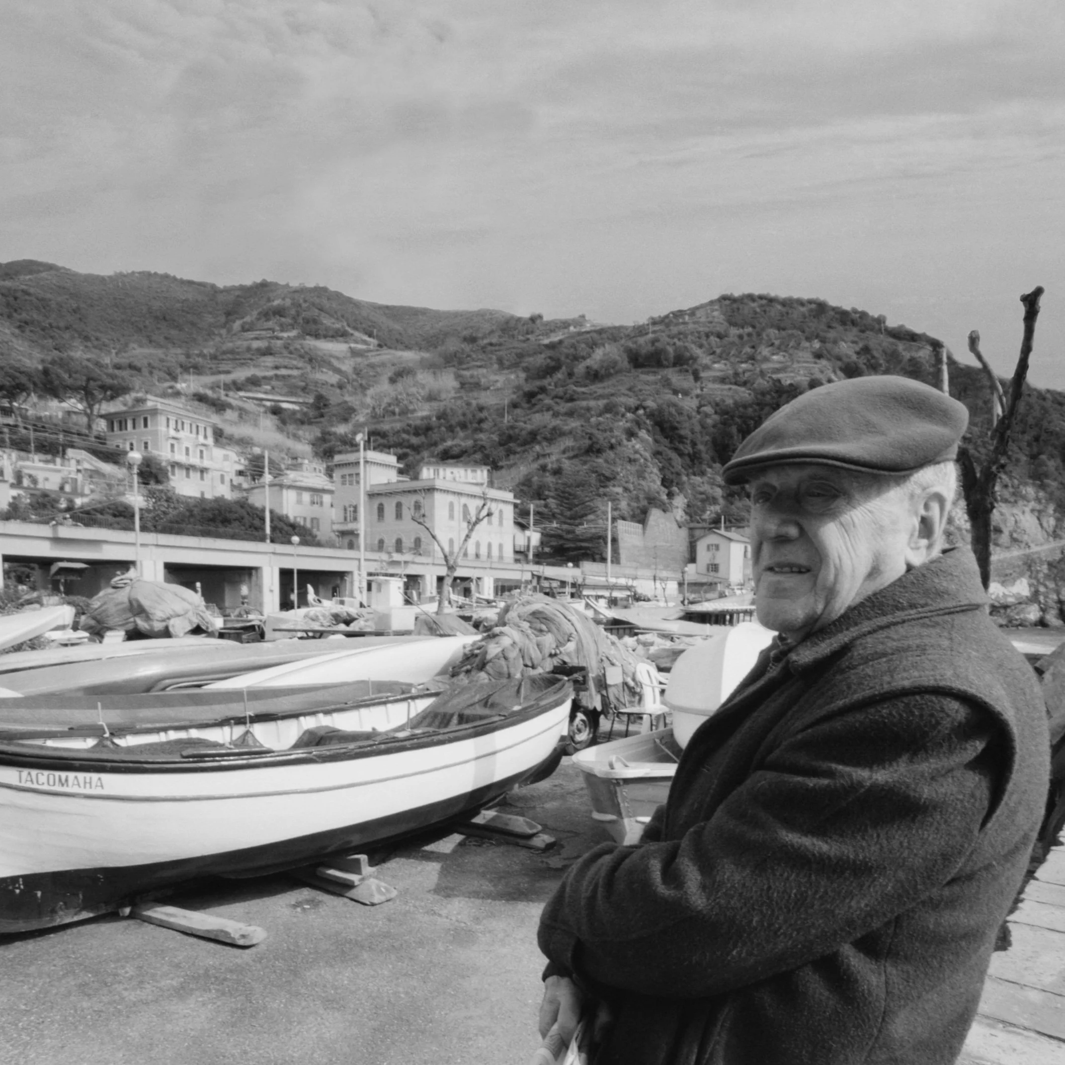 An elderly man wearing a flat cap and coat standing near boats docked at a harbor with hillside houses in the background.