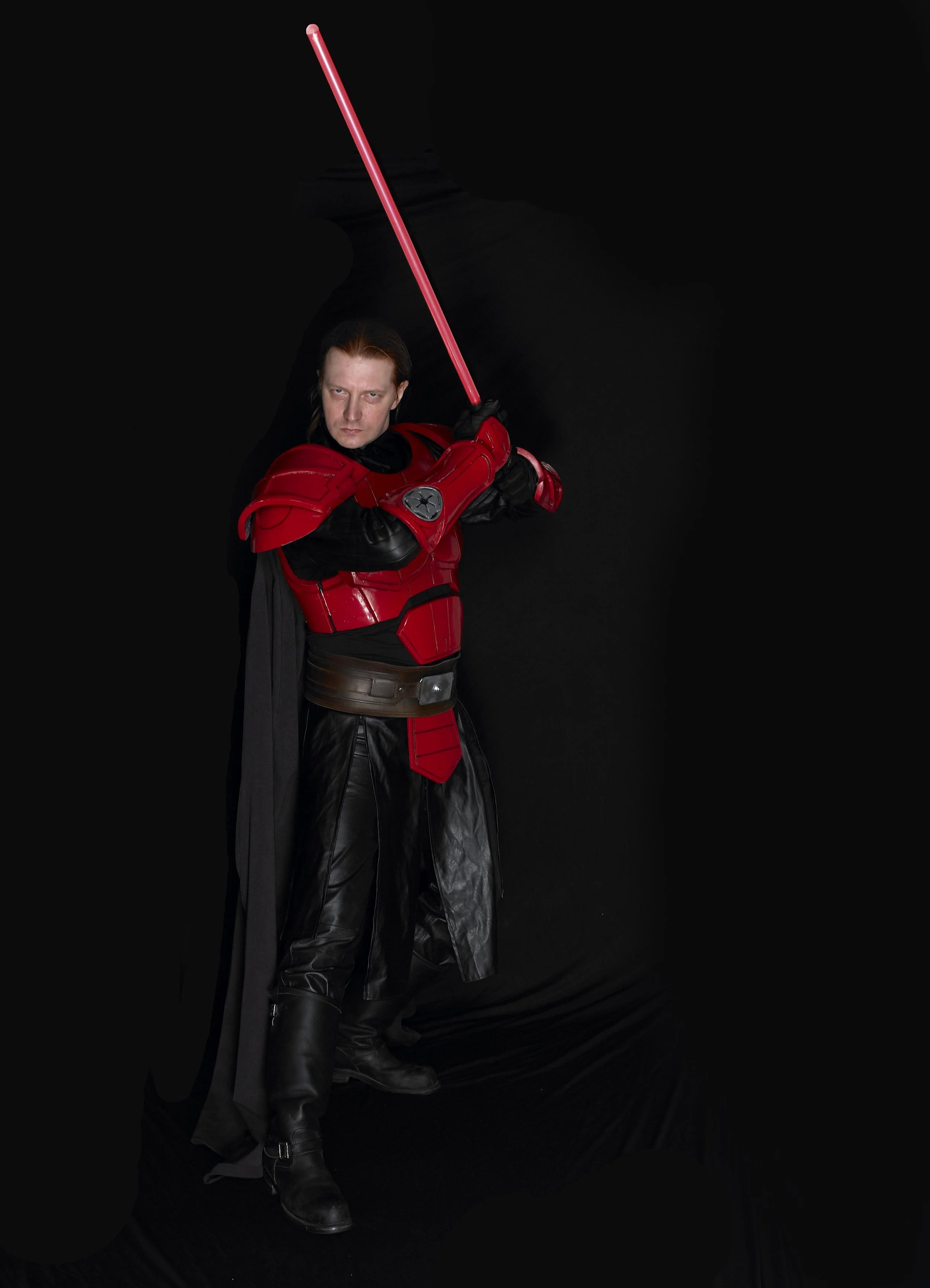 Person dressed as a Star Wars character, wearing black leather pants, red armor, and holding a red lightsaber, standing against a black background.
