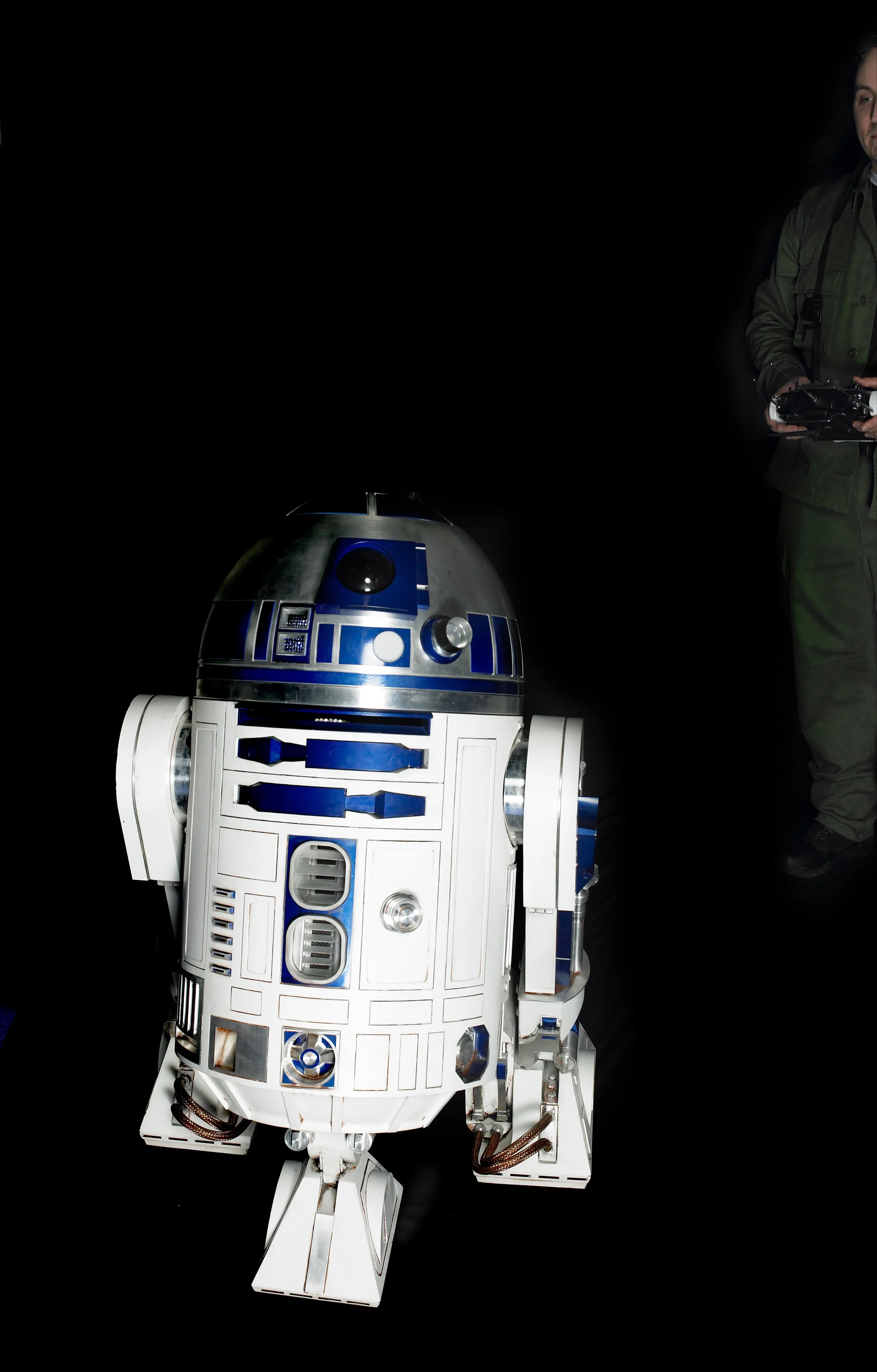 A detailed replica of the Star Wars character R2-D2 in white, blue, and metallic colors, with a person holding a remote control device partially visible on the right side of the dark background.