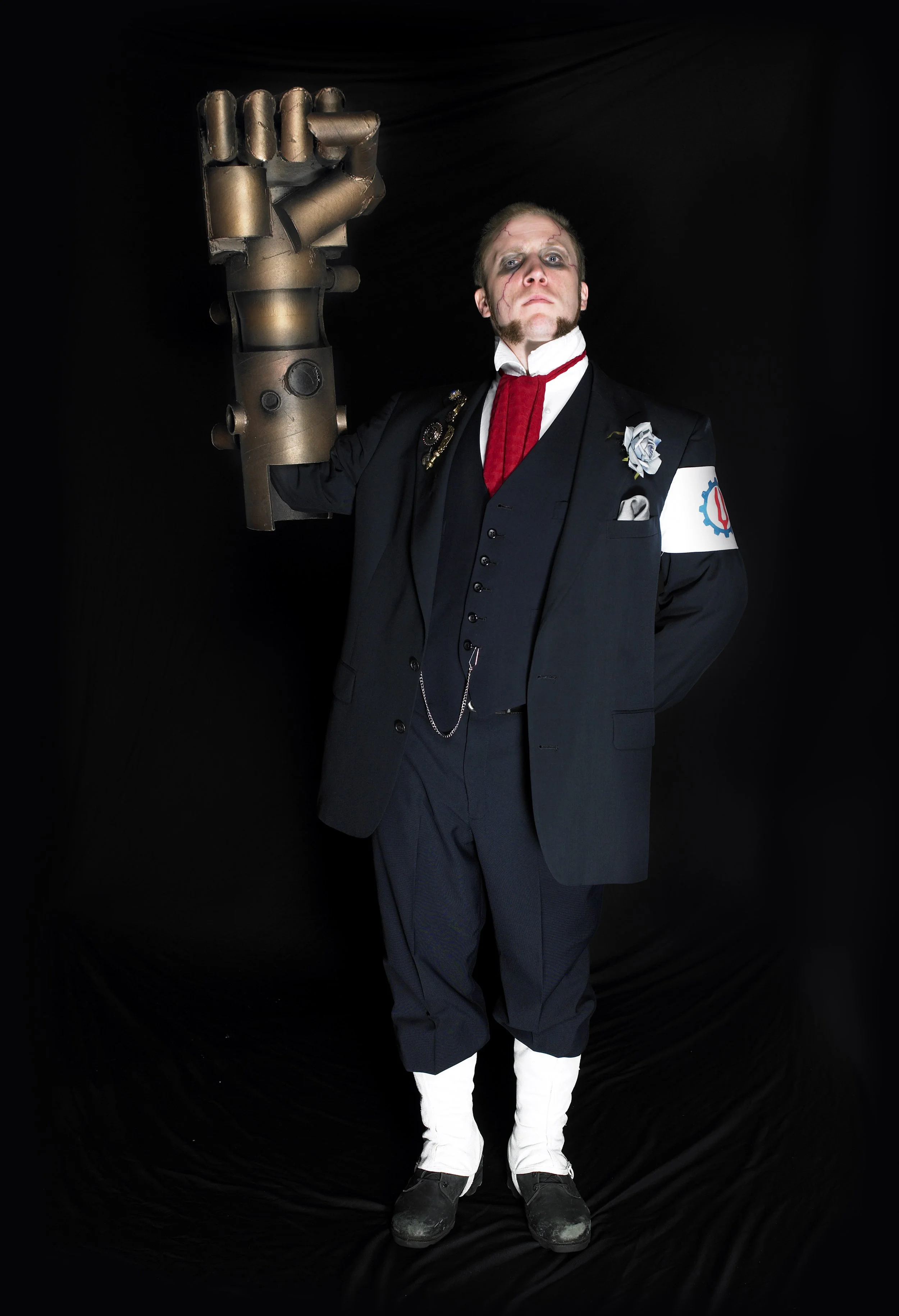 Person dressed in a steampunk-inspired costume with a dark suit, white shirt, red tie, and white stockings, holding a large mechanical fist prop against a black background.