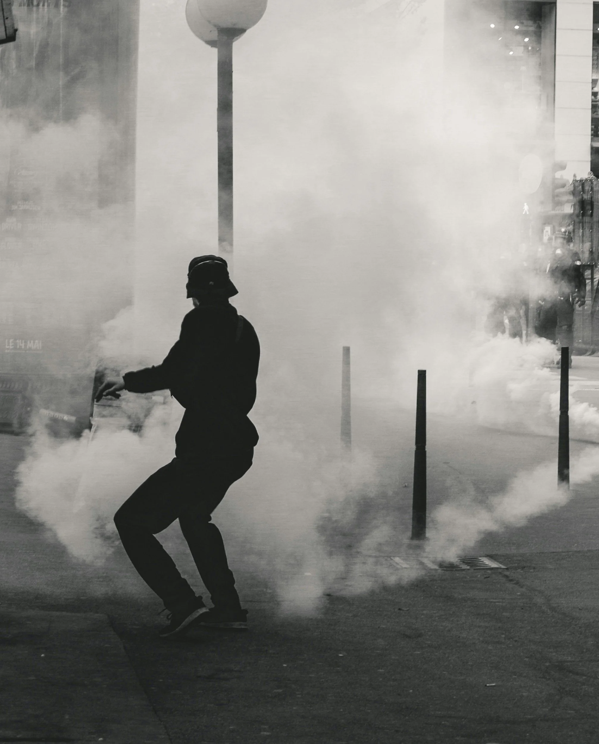A person wearing a helmet and dark clothing stands amid smoke or tear gas on a city street.