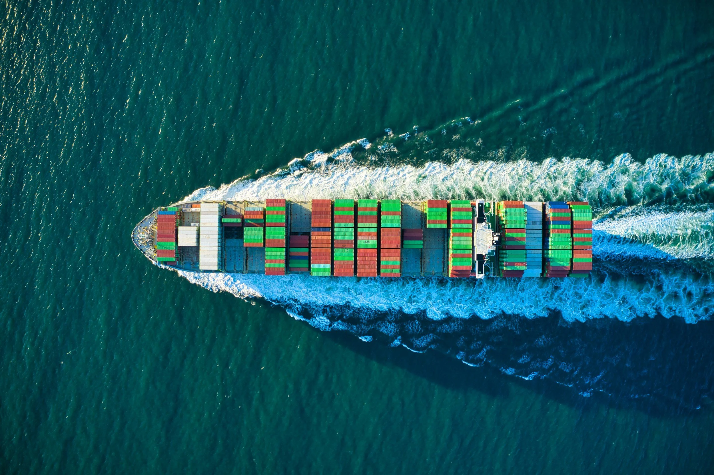Aerial view of a cargo ship carrying multicolored containers moving through the ocean, leaving a wake behind.